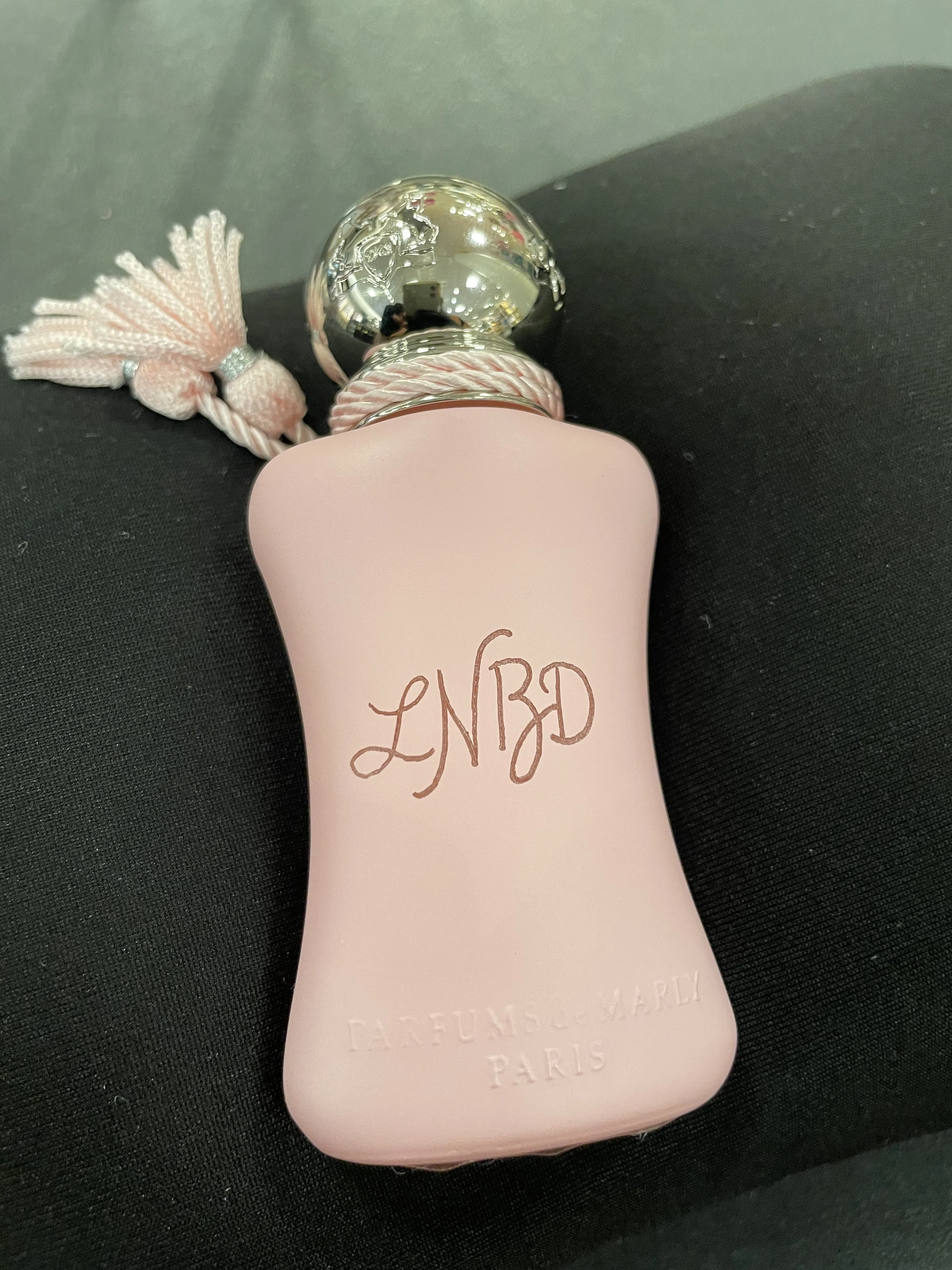 A pale pink bottle with a silver cap and a pink tassel, lying on a black surface.