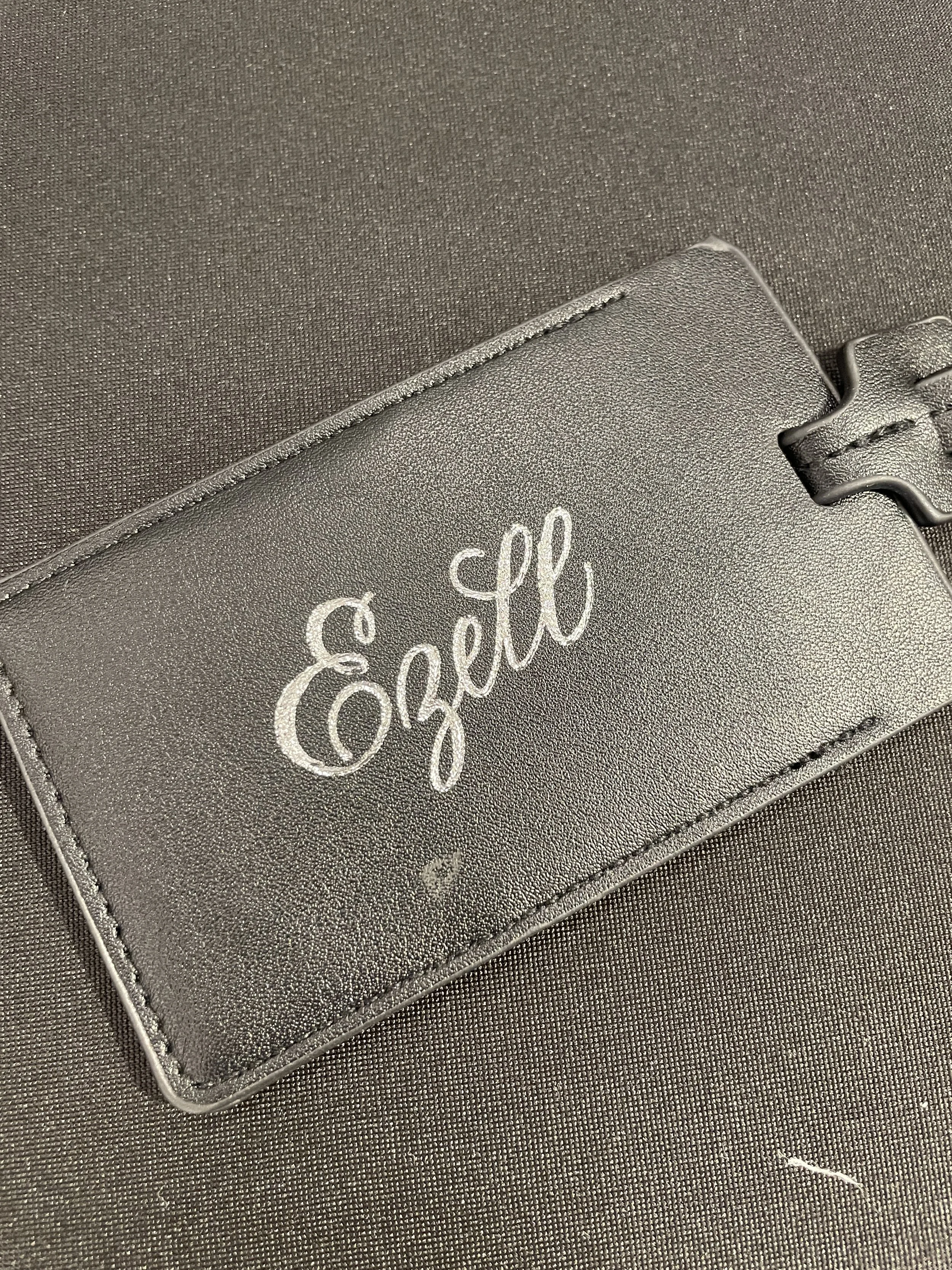 A gray leather wallet with the word 'Ezeke' written in white cursive on the front, placed on a dark textured surface.