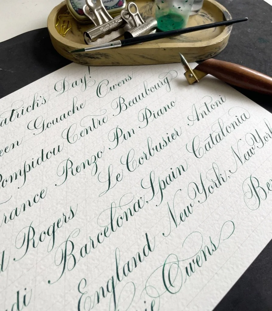 A sheet of paper with elegant calligraphy listing city names, placed on a black surface. A wooden-handled calligraphy pen, a metallic nib, and a small tray with paint, a brush, and metallic stampers are nearby.