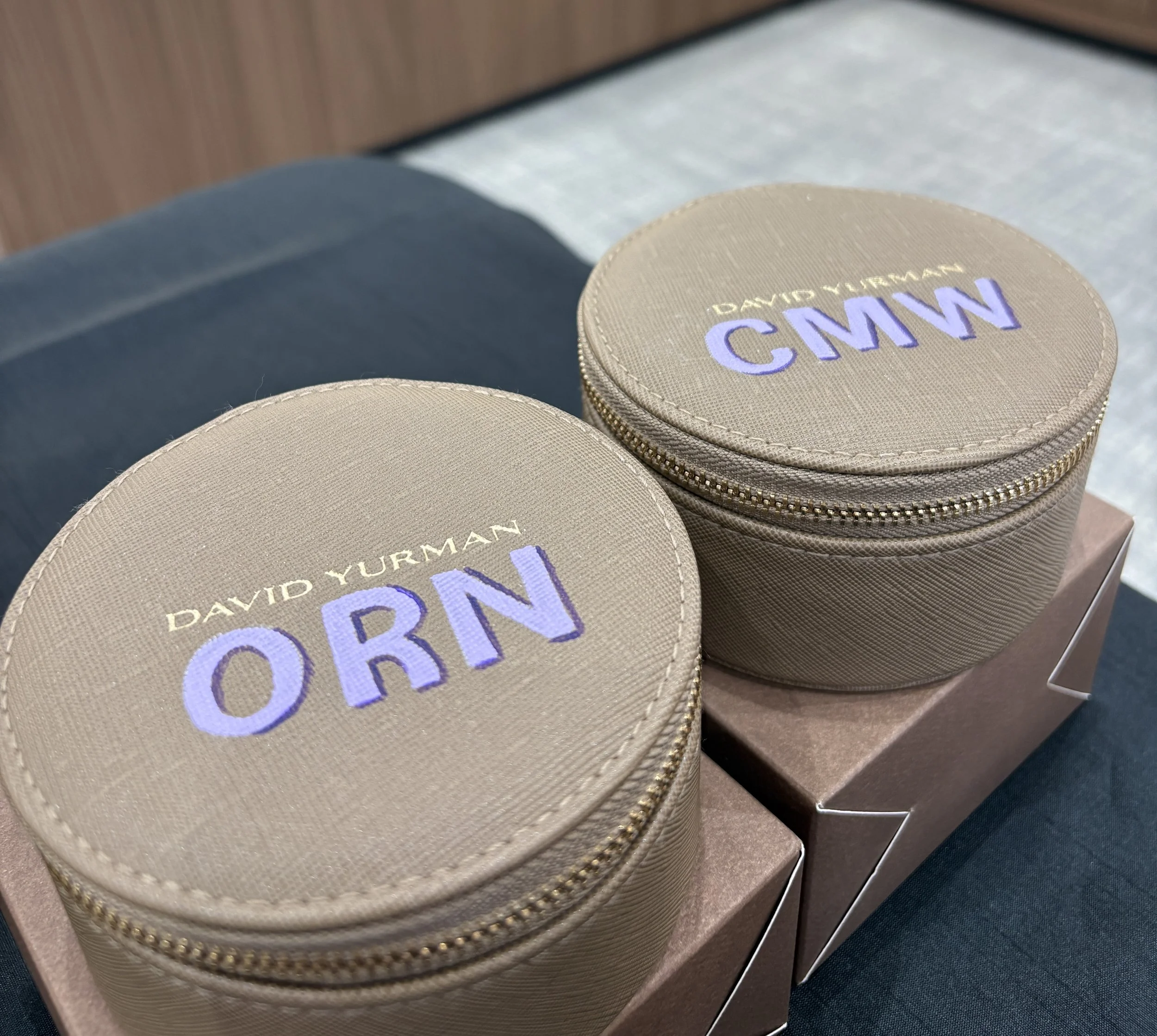 Two beige round cases with gold zippers, labeled 'David Yurman CMW' and 'ORN' in purple and gold lettering, placed on brown boxes.