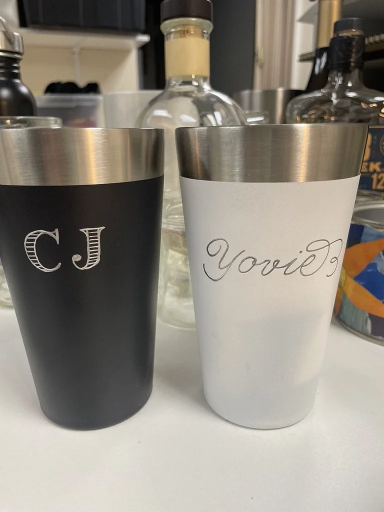 Two insulated tumblers, one black with initials 'CJ' and one white with the word 'Yours' engraved in cursive, placed on a white surface.