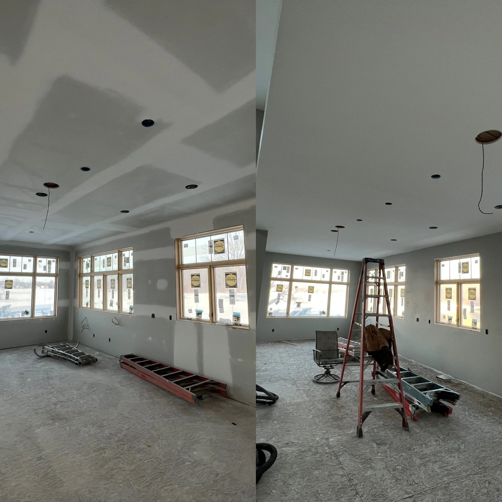 Interior view of a room under construction with drywall, windows, and construction tools, including ladders and a chair, in a well-lit space.