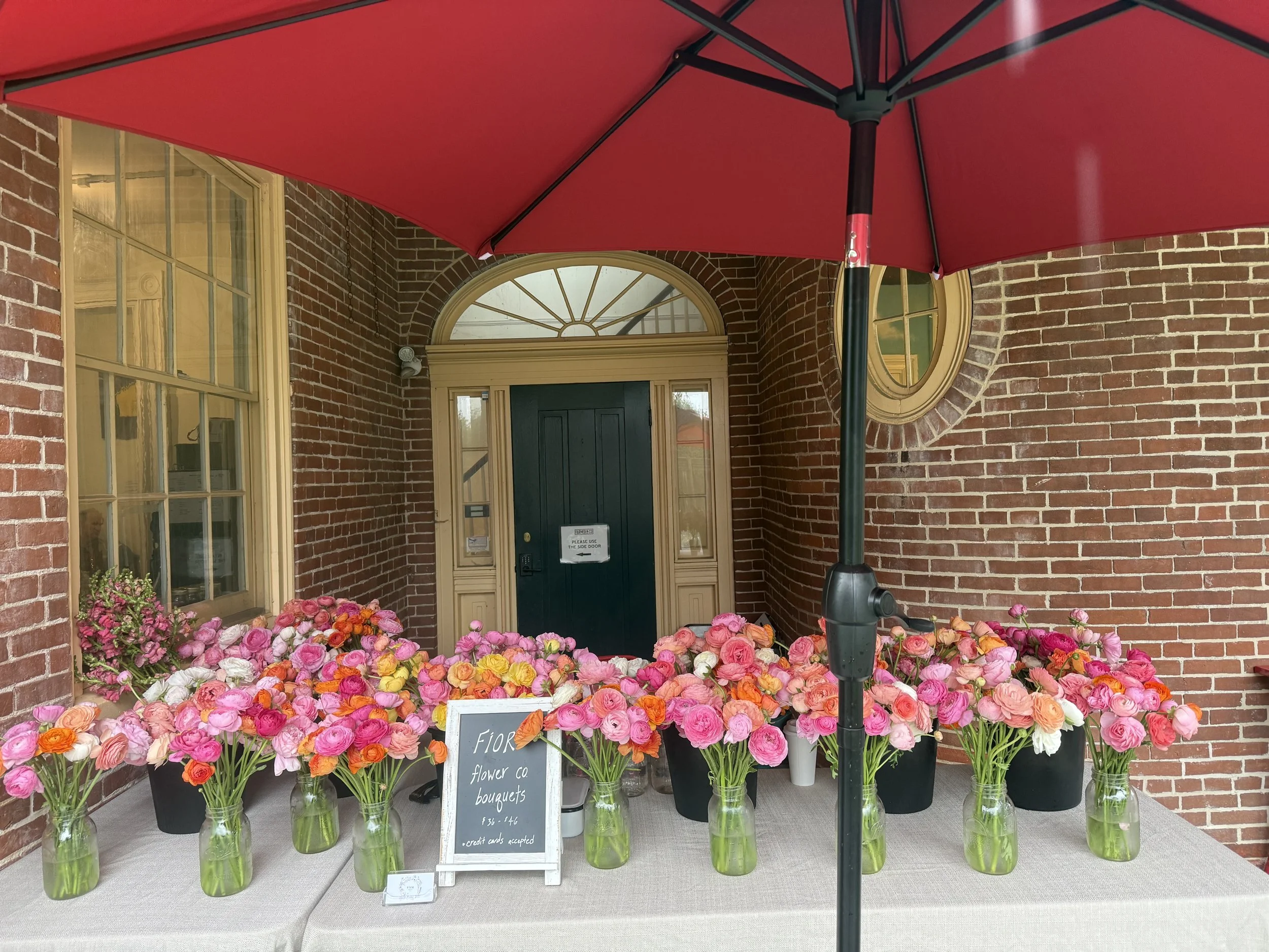 Elegant, abundant, whimsical vases of multi-colored ranunculus flowers set up on a table at a pop-up market at Farmer & Co. in Unionville, PA.
