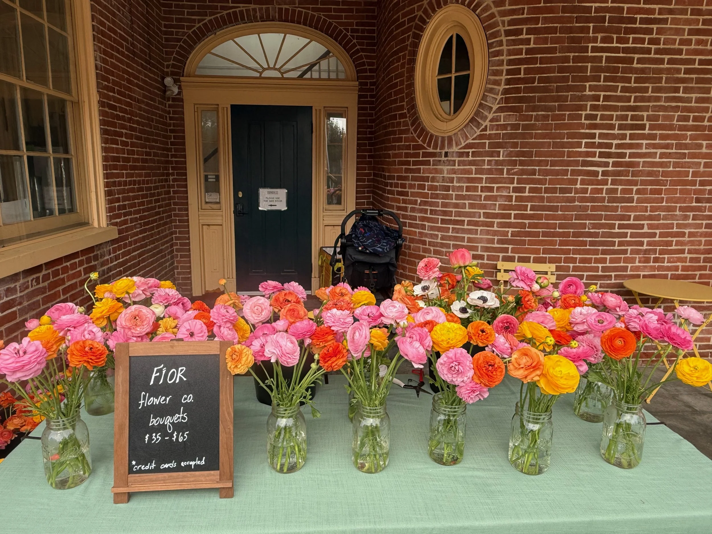 Elegant, abundant, whimsical vases of multi-colored ranunculus flowers set up on a table at a pop-up market at Farmer & Co. in Unionville, PA. 
