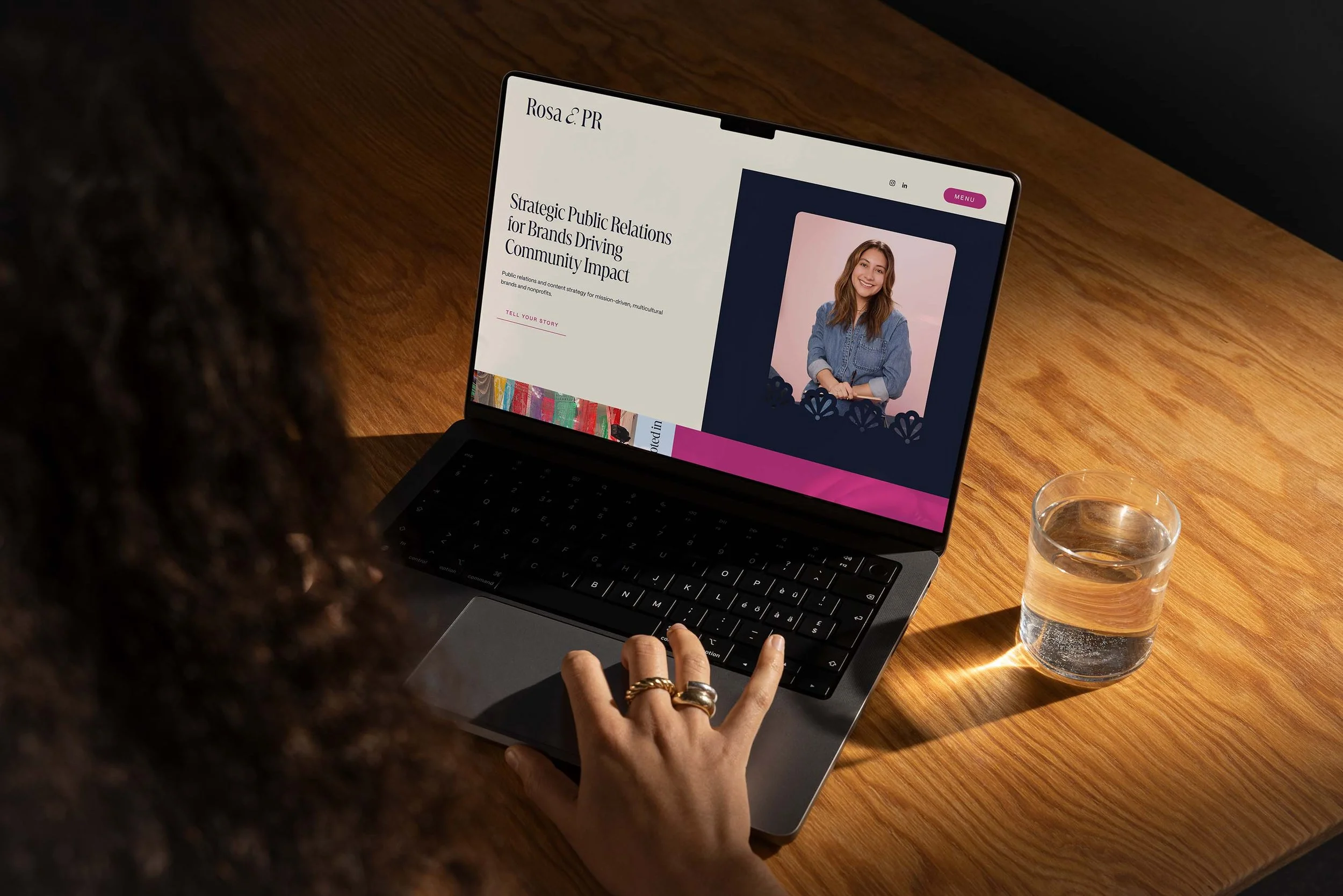 Person with curly hair using a laptop on a wooden table, with a glass of water nearby and a website open showing a woman with long hair and a blue shirt.