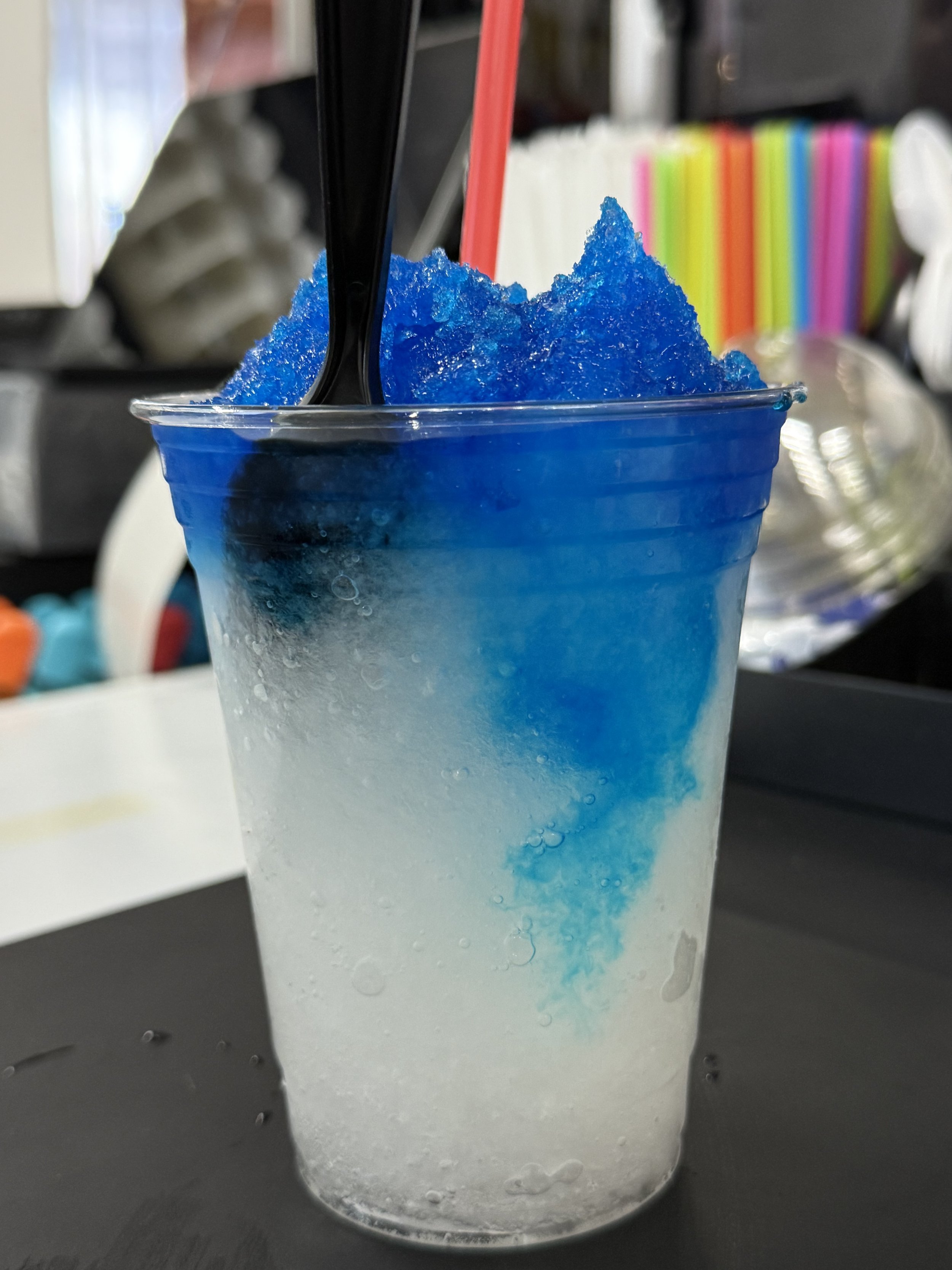 A clear plastic cup filled with blue and white syrup and shaved ice, with a red straw and black spoon inserted, on a black surface.