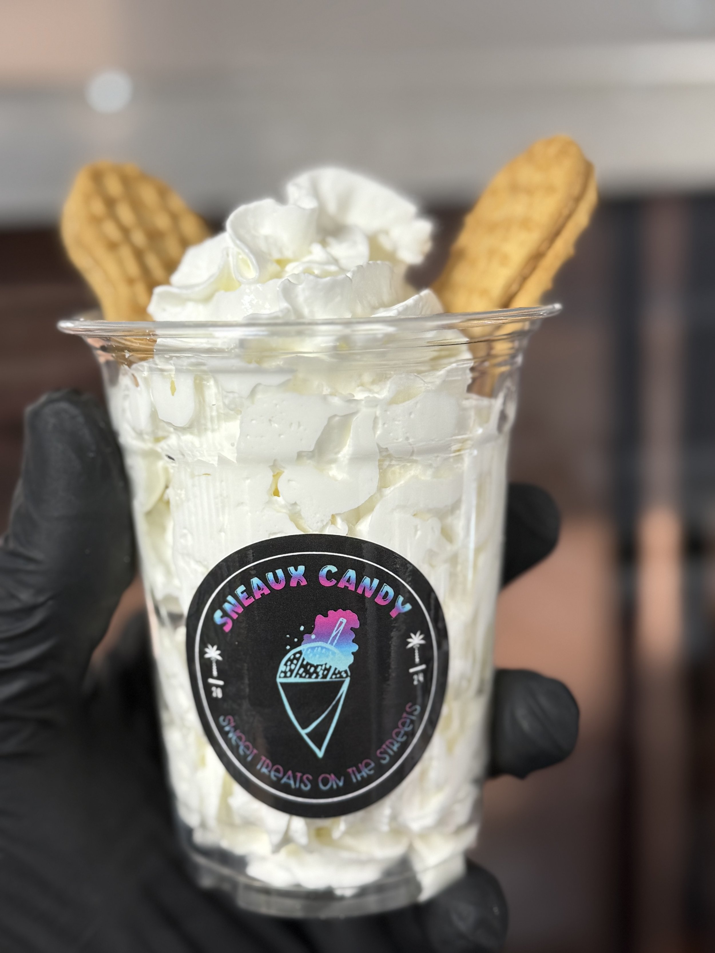 A clear plastic cup filled with whipped cream and two cookies, labeled 'Sneaux Candy' with a slogan 'Sweet Treats on the Streets'.