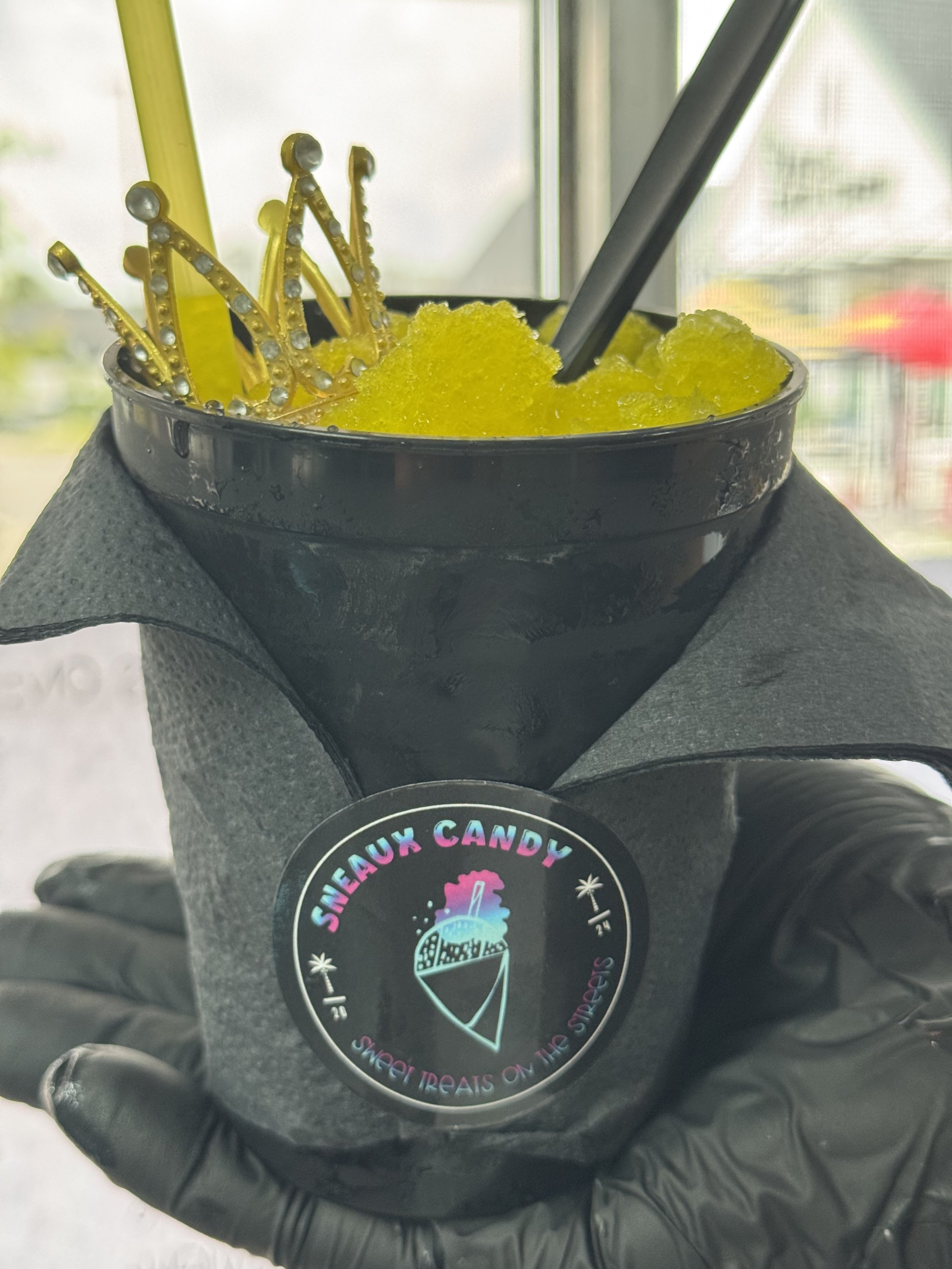 Black cup filled with yellow shaved ice topped with a yellow crown decoration and a black straw. The cup has a sticker that says 'SNEAUX CANDY' with an ice cream cone graphic, and the phrase 'SWEET TREATS ON THE STREET'S'.