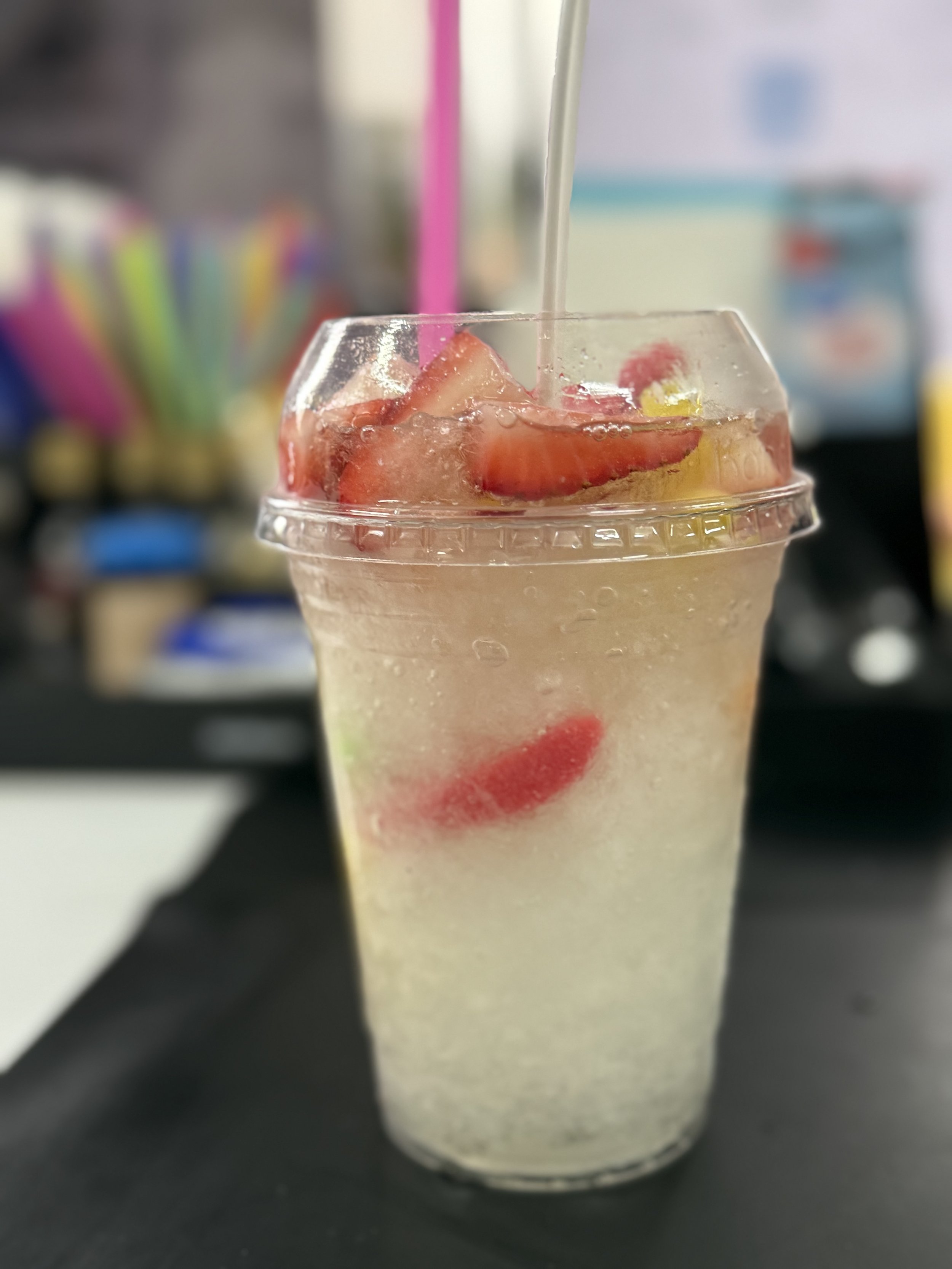 A clear plastic cup filled with Wedding Cake Syrup, shaved ice, gummy worms and sliced strawberries, with a straw & topped with white chocolate.