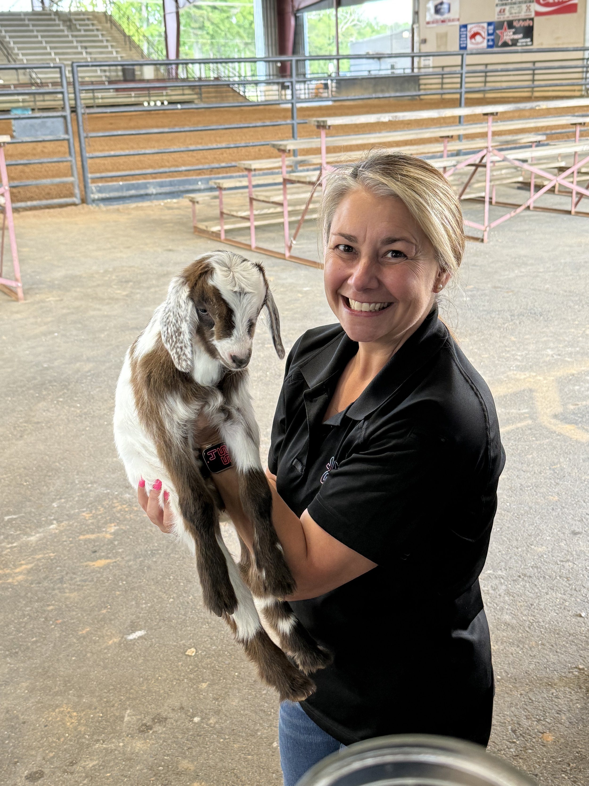 A woman smiling and holding a brown and white goat kid inside a barn or covered area with bleachers in the background.