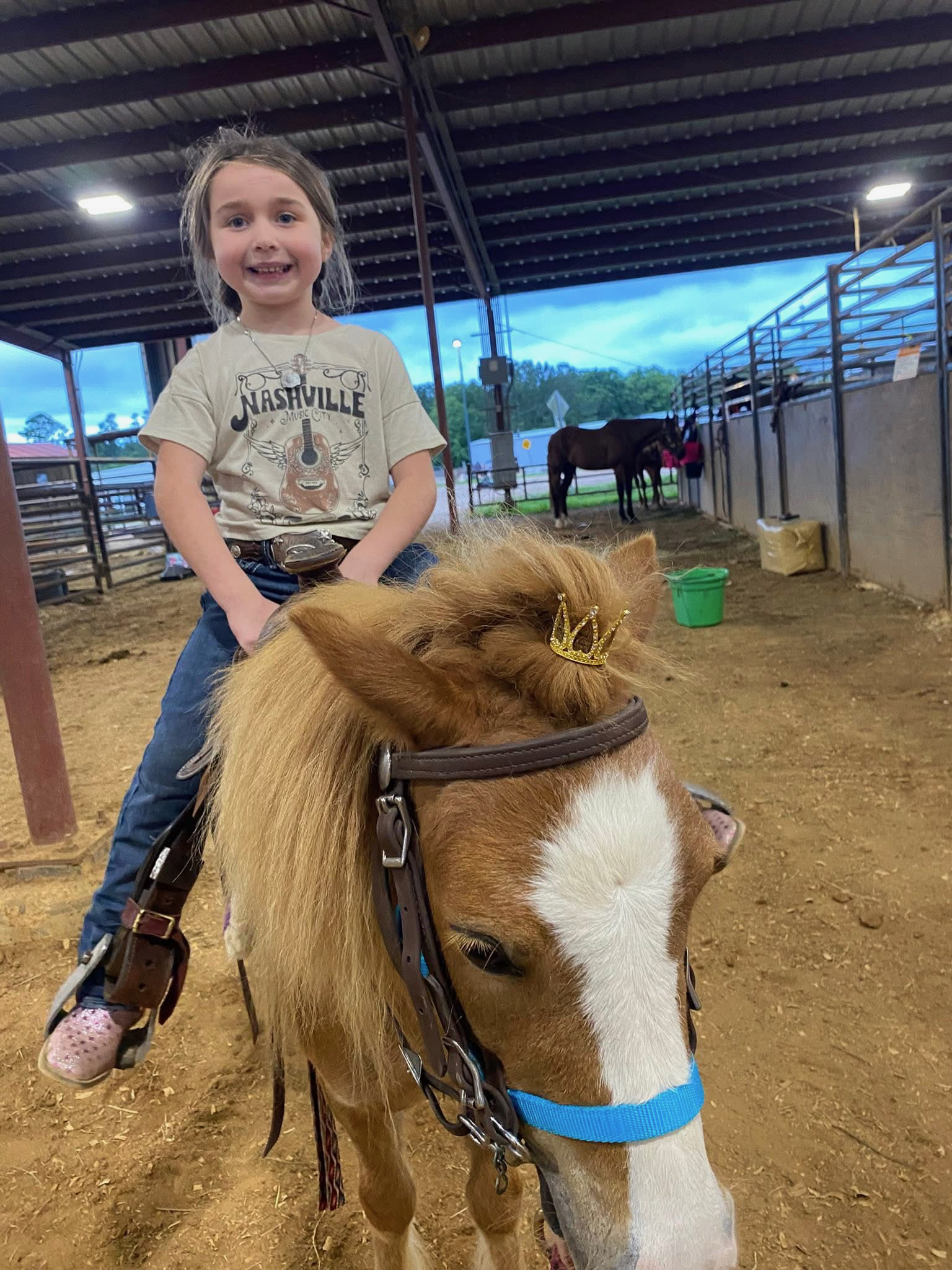 Young girl with long hair, smiling and wearing a Nashville t-shirt, sitting on a small horse with a gold crown on its head in an indoor equestrian center, with other horses and visitors in the background.
