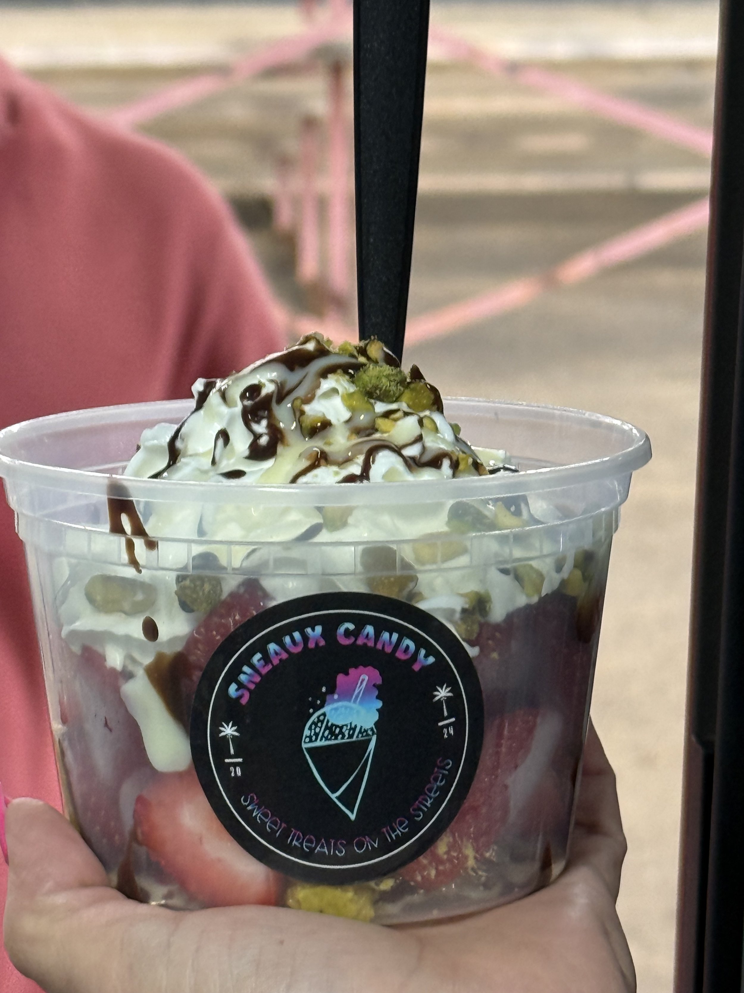 A clear plastic cup filled with strawberries, topped with whipped cream, chocolate syrup, and chopped pistachios, with a black straw. The cup has a sticker that says 'SHEAUX CANDY' and 'Sweet Treats on the Streets' with an ice cream cone logo.