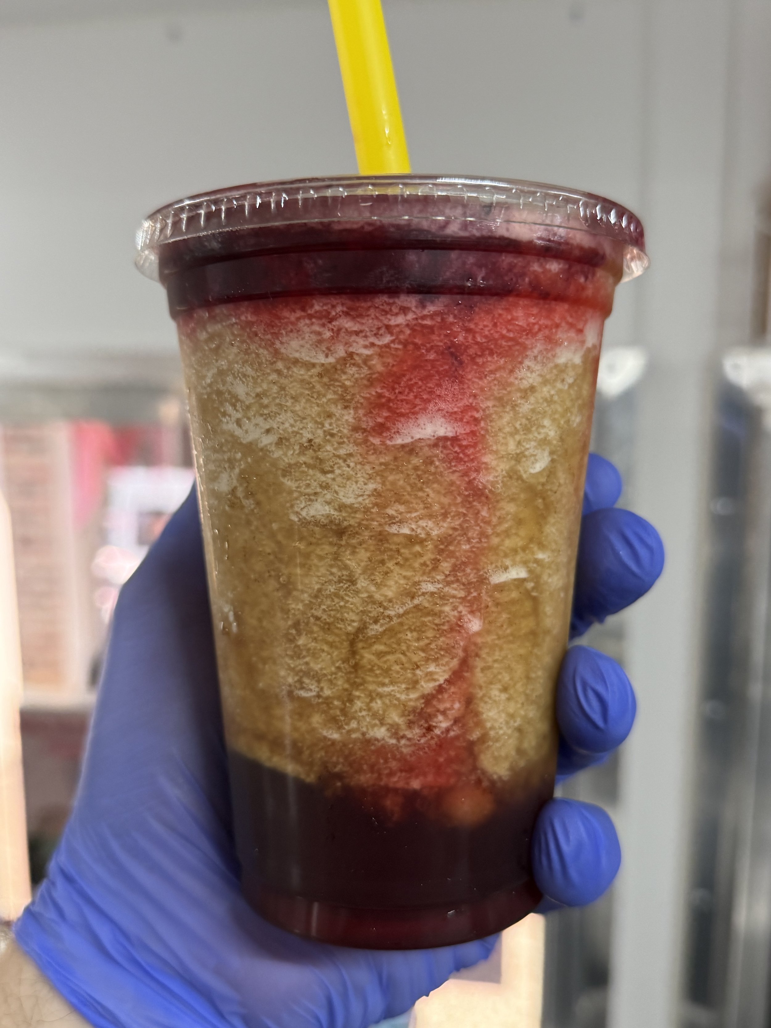 A cold beverage in a clear plastic cup with a yellow straw, showing dark red and yellowish layers, held by a person wearing a blue glove.