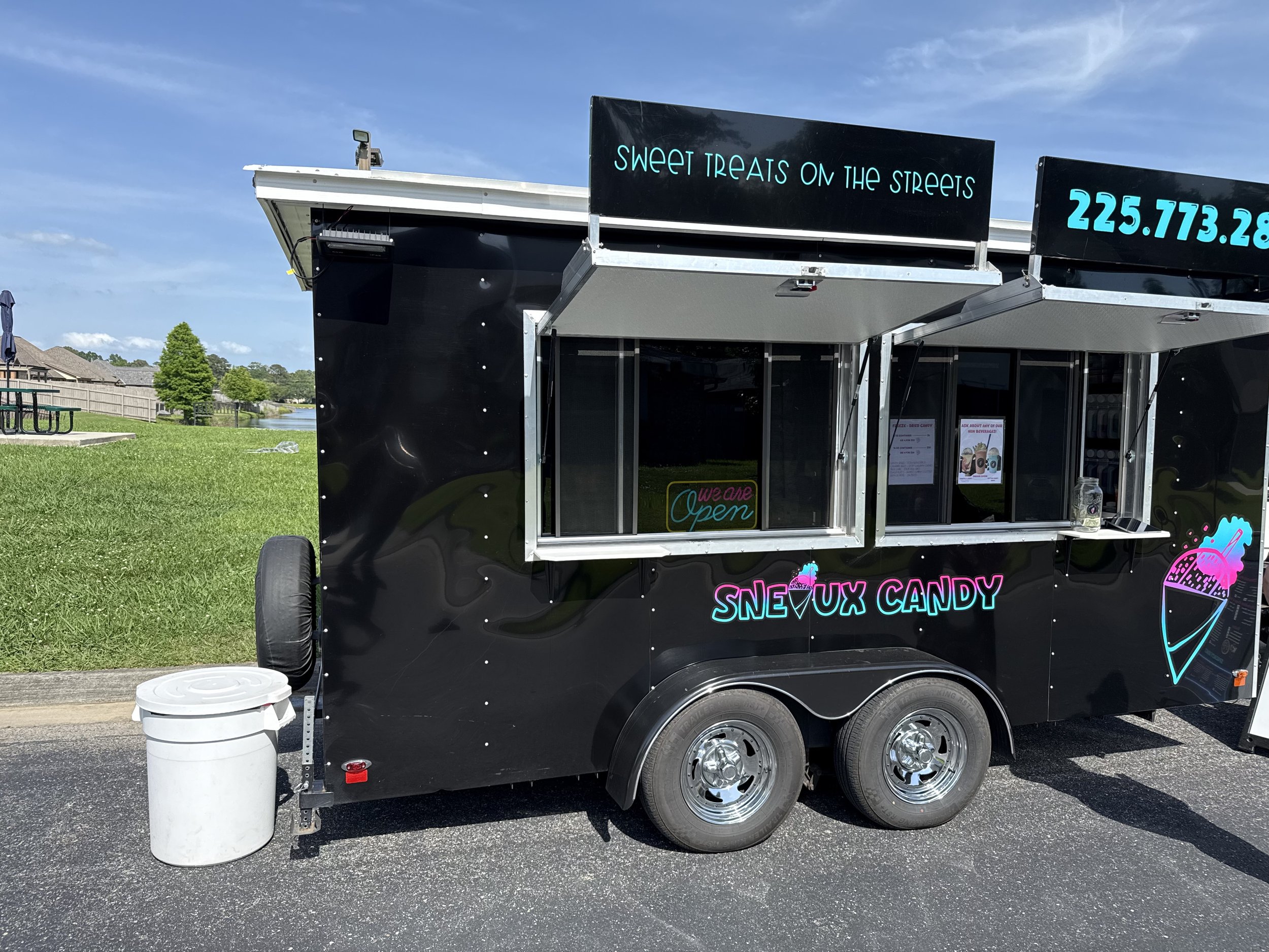 Black candy food truck with neon signs advertising snow cone candy and sweet treats on the streets, parked on a paved road with a grassy park, trees, and a pond in the background.
