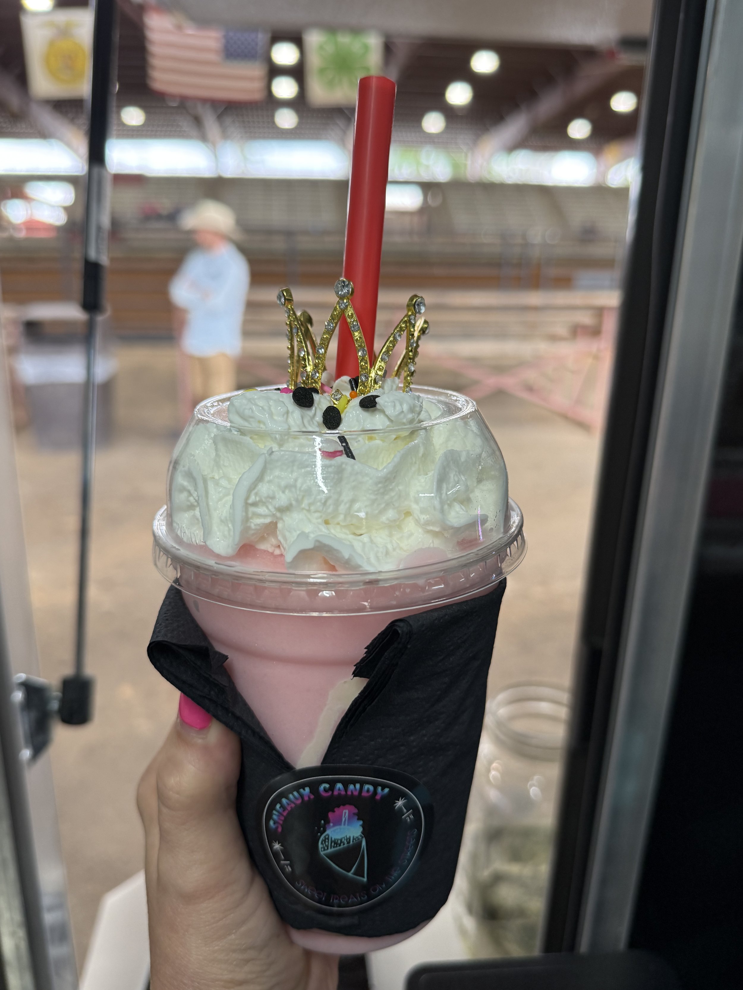 A pink milkshake topped with whipped cream, decorated with a small gold princess crown, colorful sprinkles, and a red straw, held in a black Sleeve Candy cup holder, in front of a blurred background of an outdoor covered area with a person in a hat.