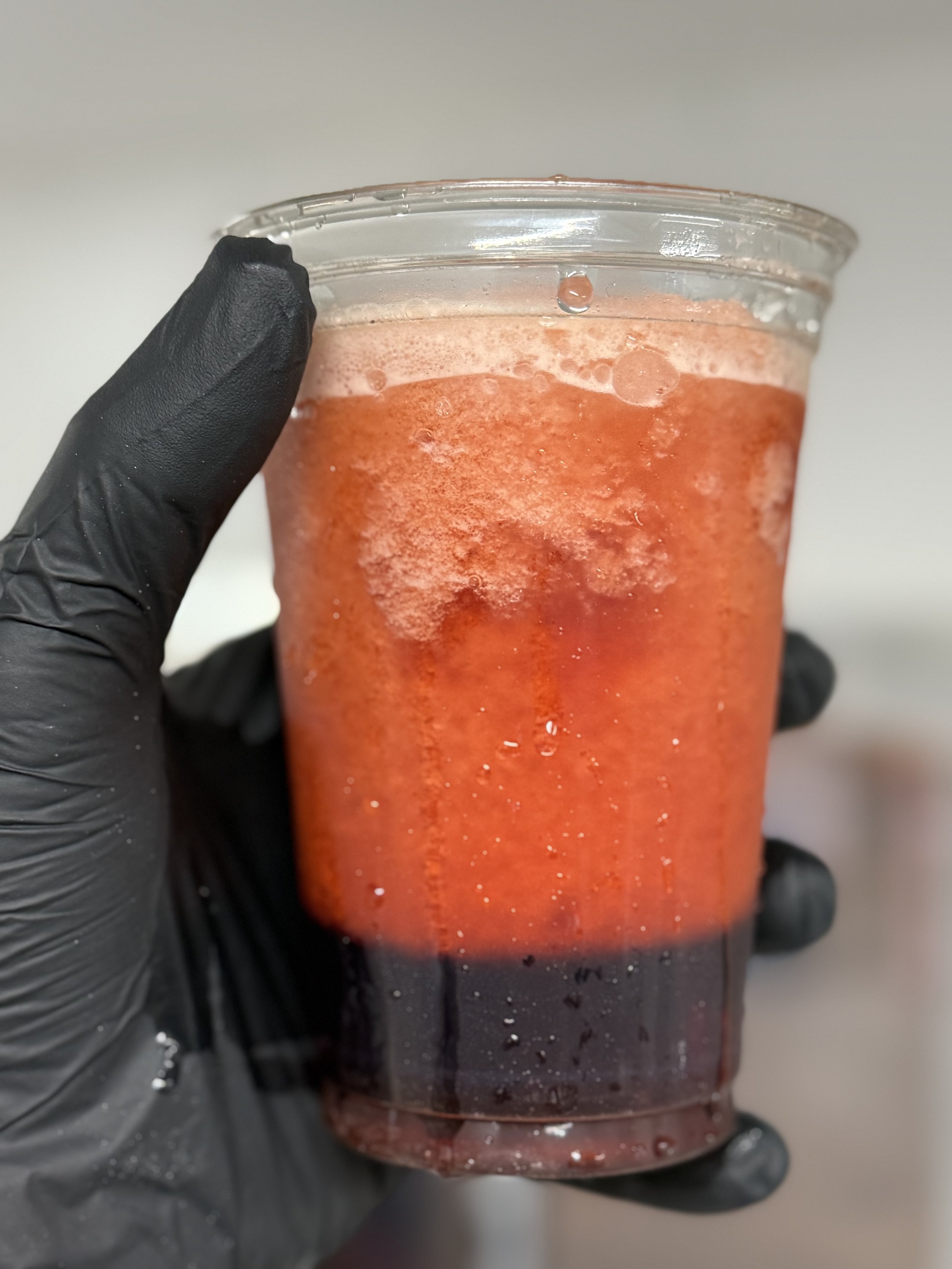A clear plastic cup with a dark red liquid, held by a hand wearing a black glove, with some foam on top.