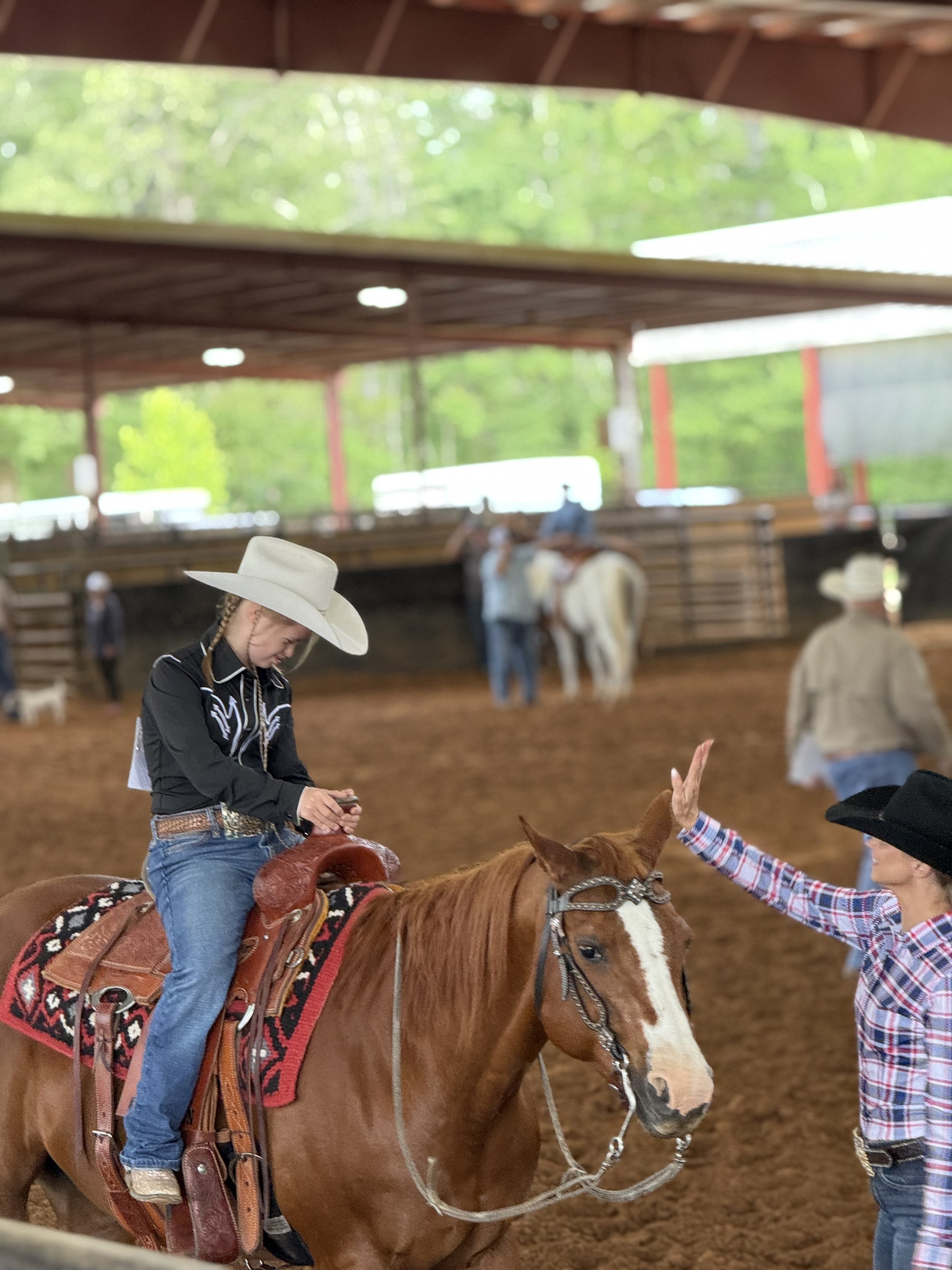 A young girl in a cowboy hat sitting on a brown horse inside a rodeo arena, while a woman in a plaid shirt and black cowboy hat high-fives her. Other riders and horses are visible in the background.