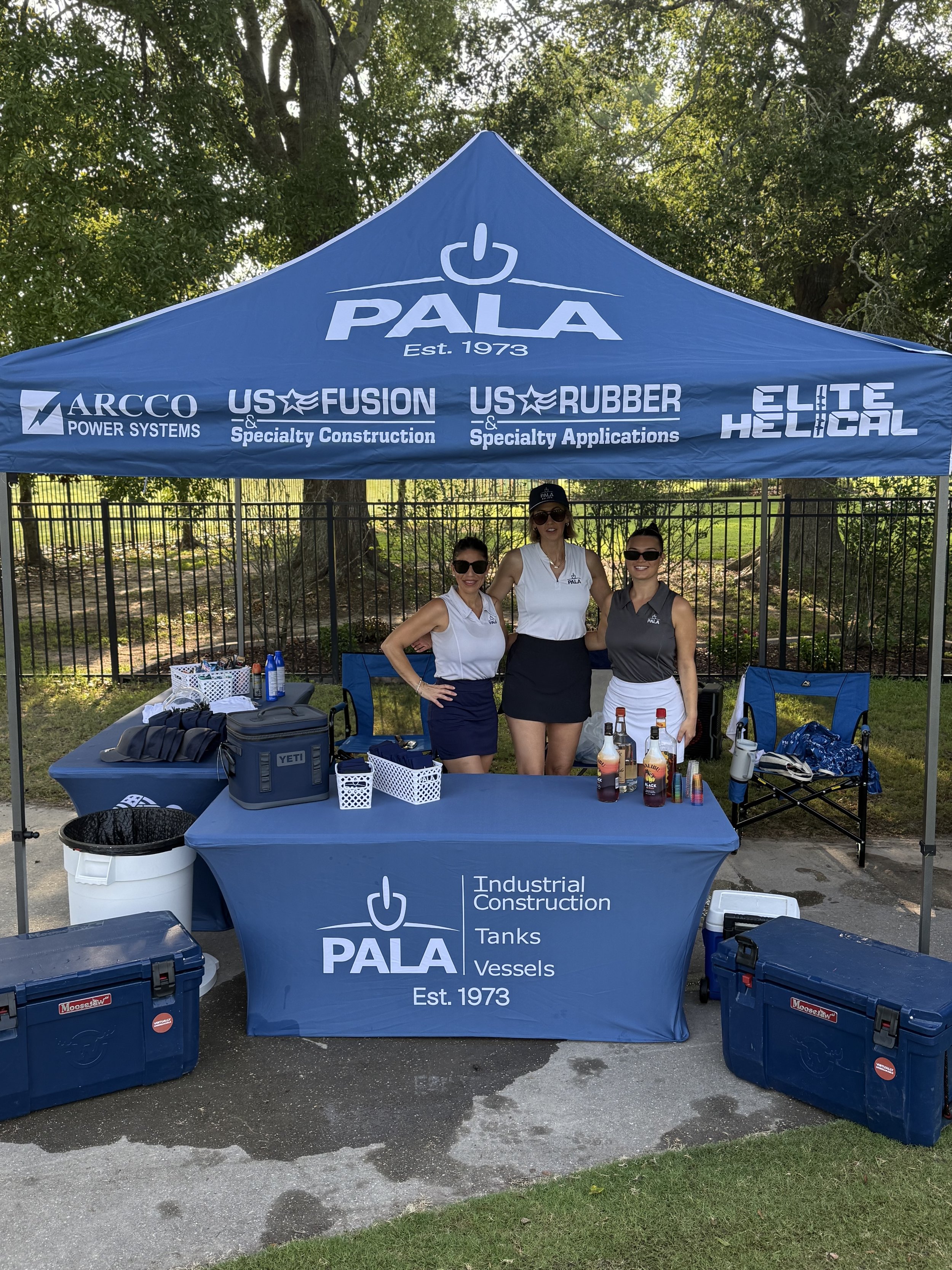 Three women standing behind a booth with a blue canopy labeled PALA, at an outdoor event. The booth displays bottles and equipment with matching tablecloths, and the women are wearing casual business attire, sunglasses, and smiling.