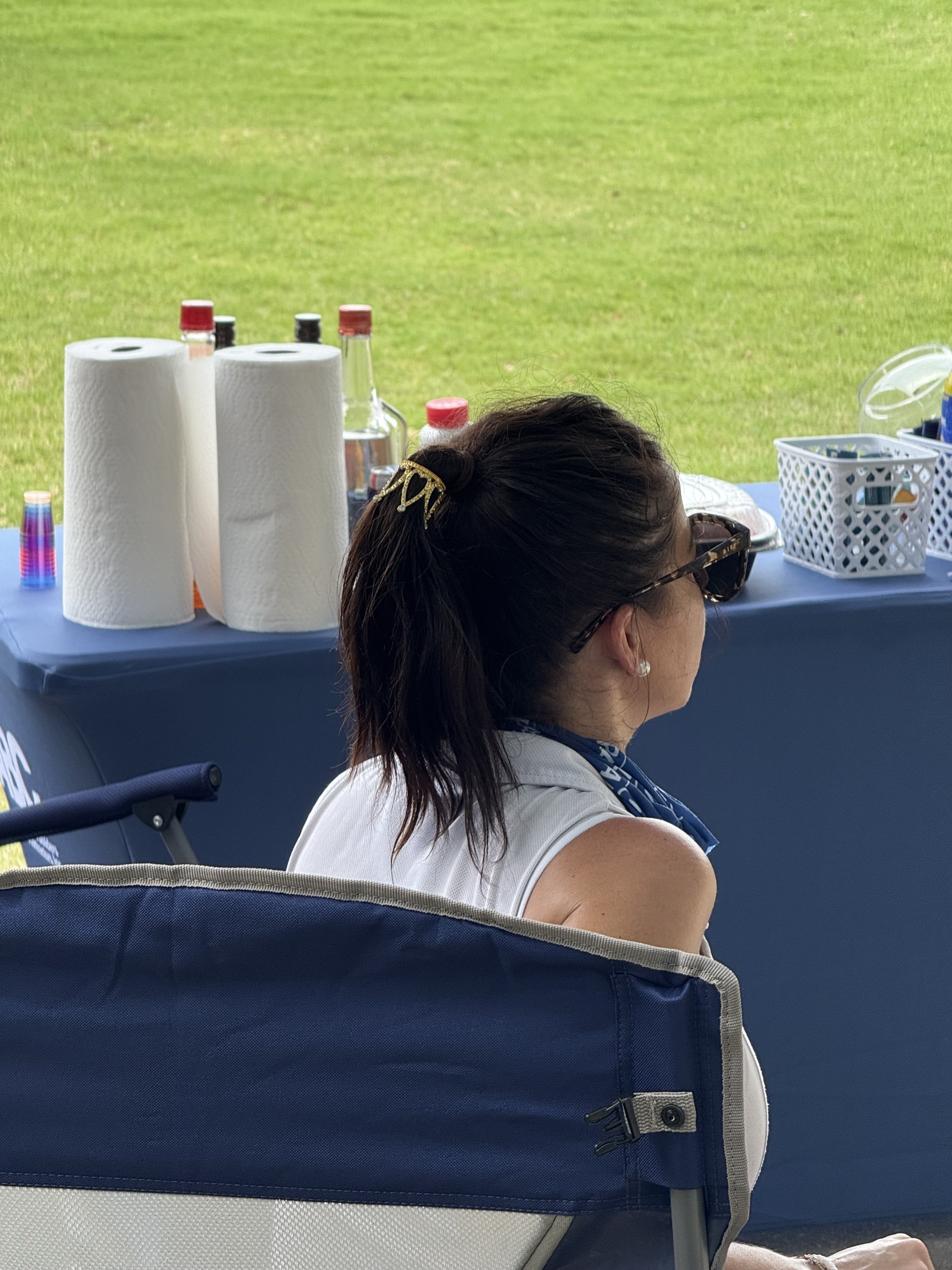 A woman with dark hair sitting at a table outdoors with a grassy field in the background. The table has paper towels, bottles, and a white basket.