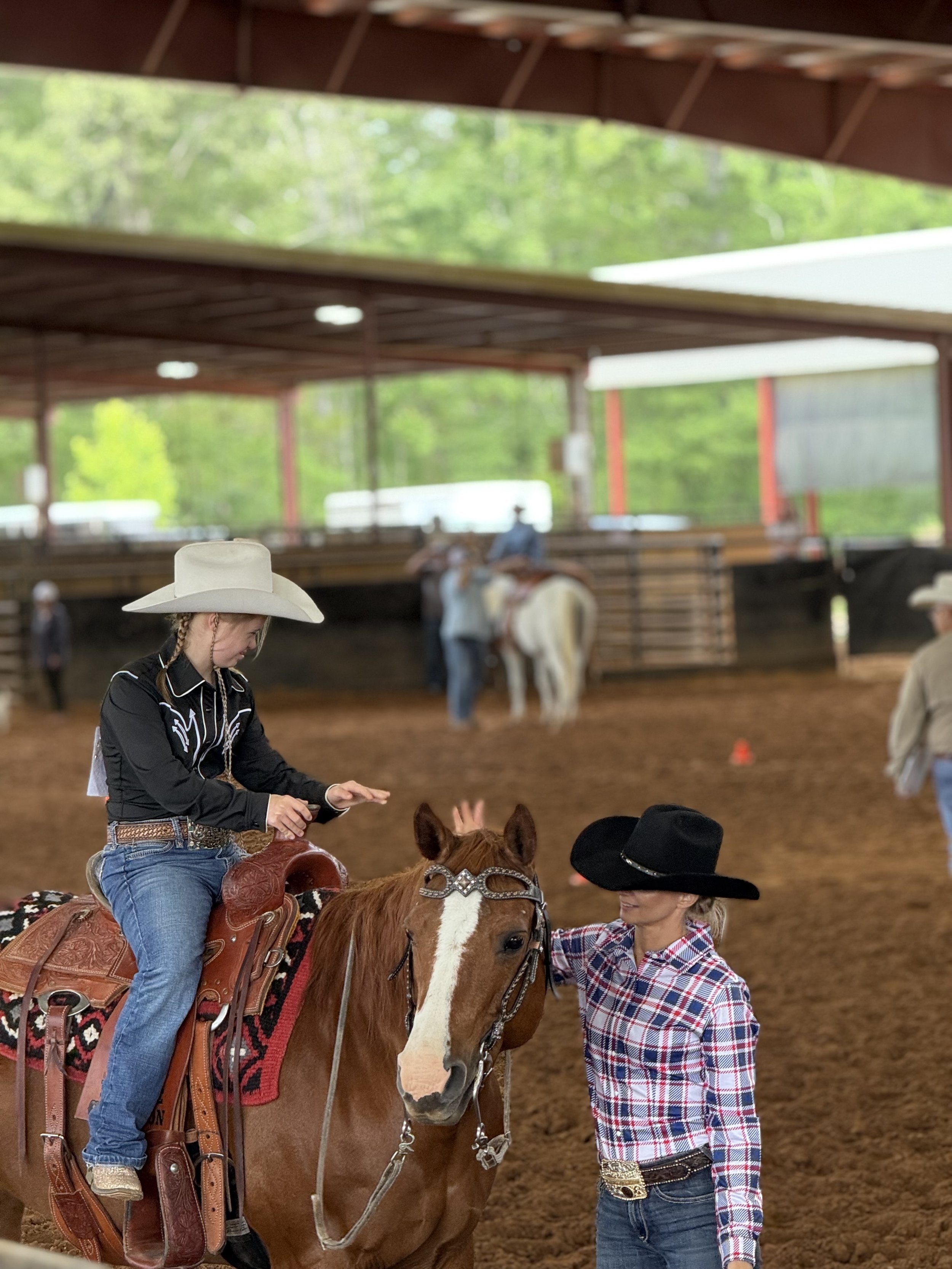 A young girl wearing a white cowboy hat and black cowboy shirt is sitting on a brown horse with a decorative saddle and bridle, while a woman in a black hat and plaid shirt stands beside them, holding the horse's reins inside an indoor riding arena.