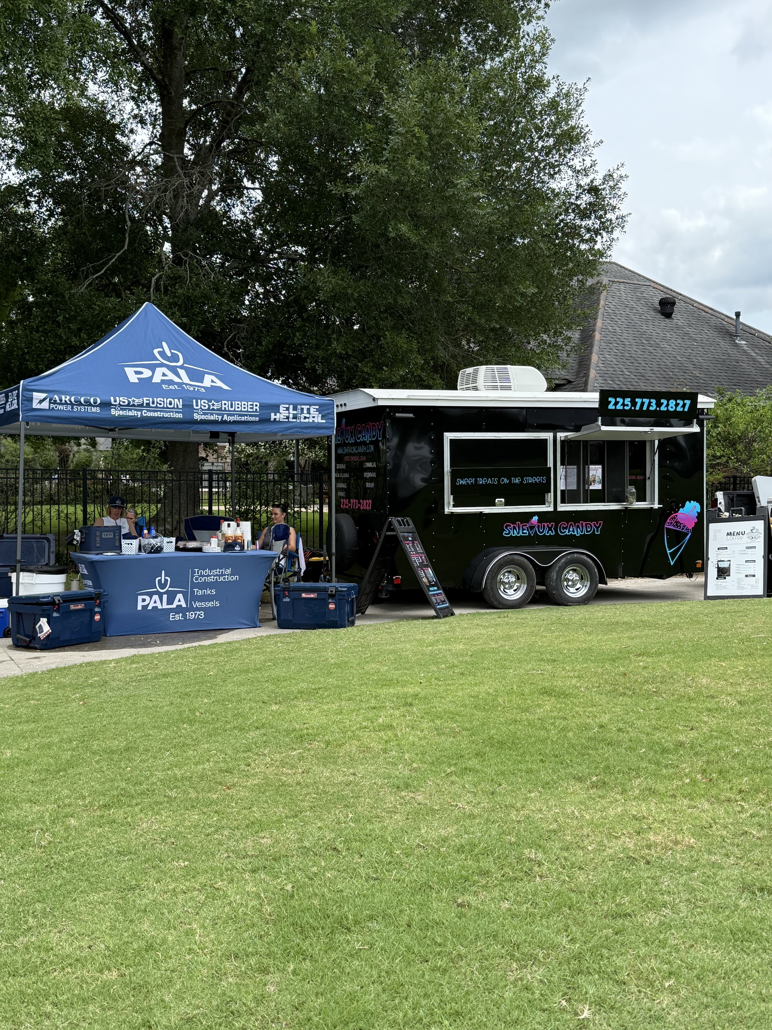 A black mobile candy stand named 'Snowx Candy' with colorful sign and menu, parked on a grassy area, with a blue tent advertising construction services nearby.