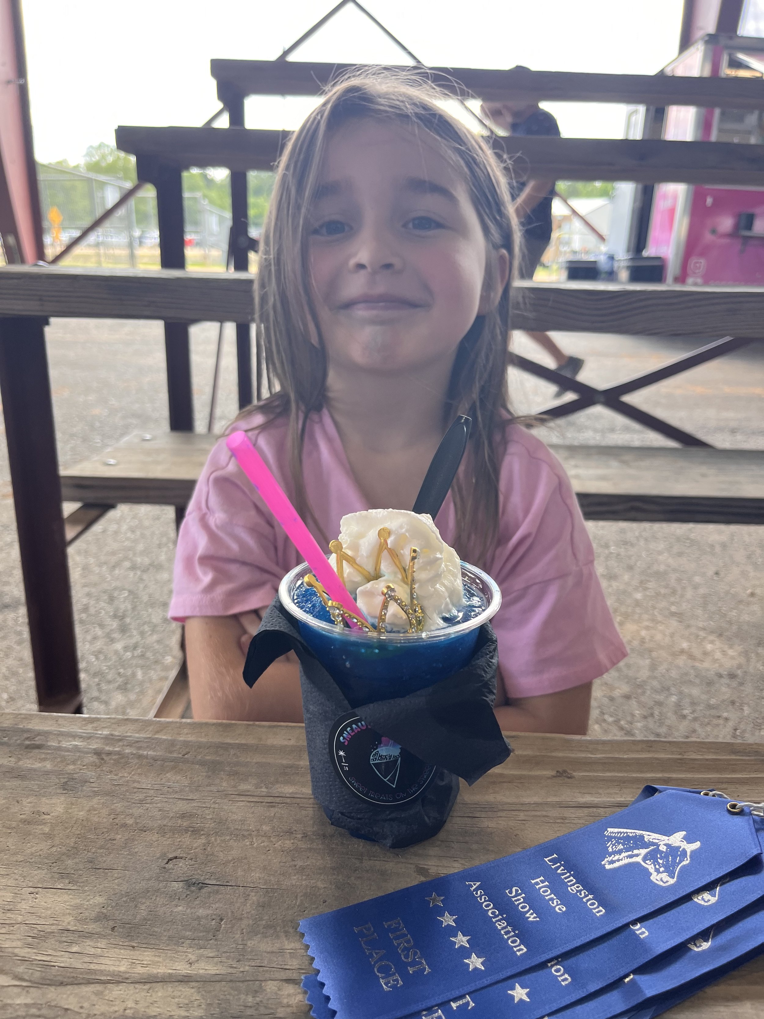 Young girl in pink shirt sitting at a wooden table with a blue frozen dessert topped with whipped cream and gold sprinkles, with blue ribbons from a horse show association beside her.