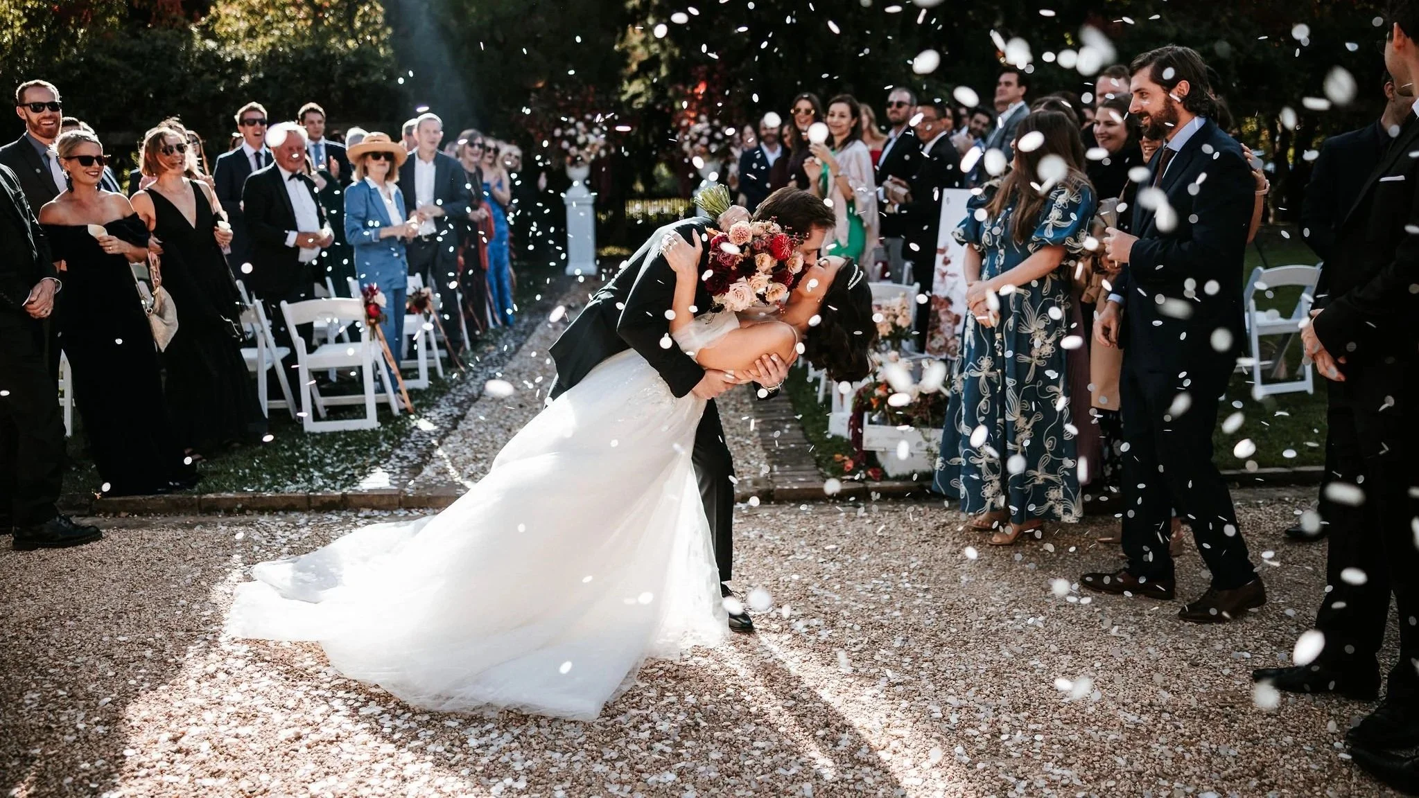 A bride and groom kiss during their outdoor wedding ceremony, surrounded by pink and white flowers.