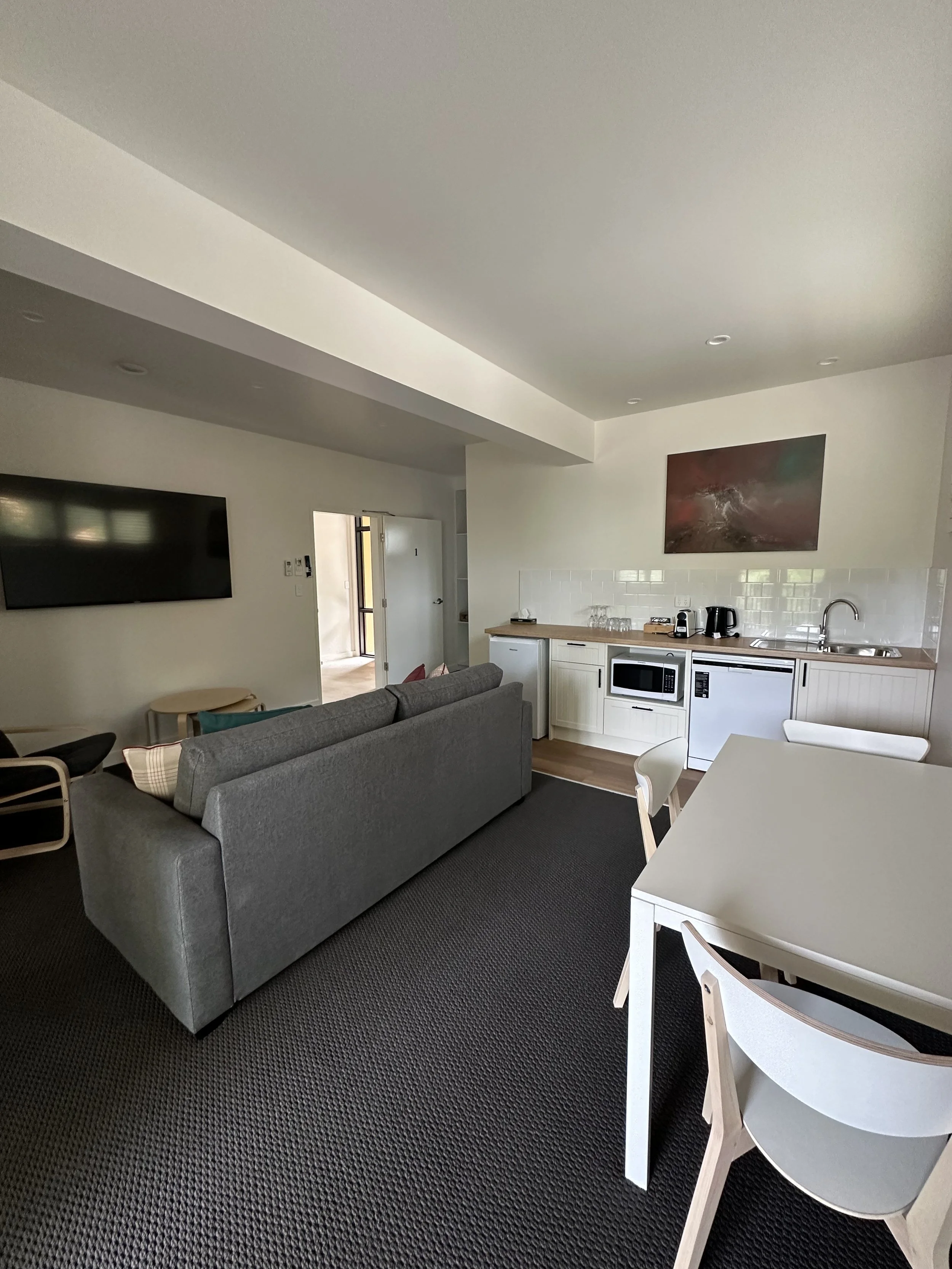 Open-concept living room and kitchenette with a flat-screen TV on the wall, a gray sofa, a white dining table with chairs, a kitchenette with a microwave, mini fridge, coffee maker, and sink, a painting on the wall, and a doorway leading outside.