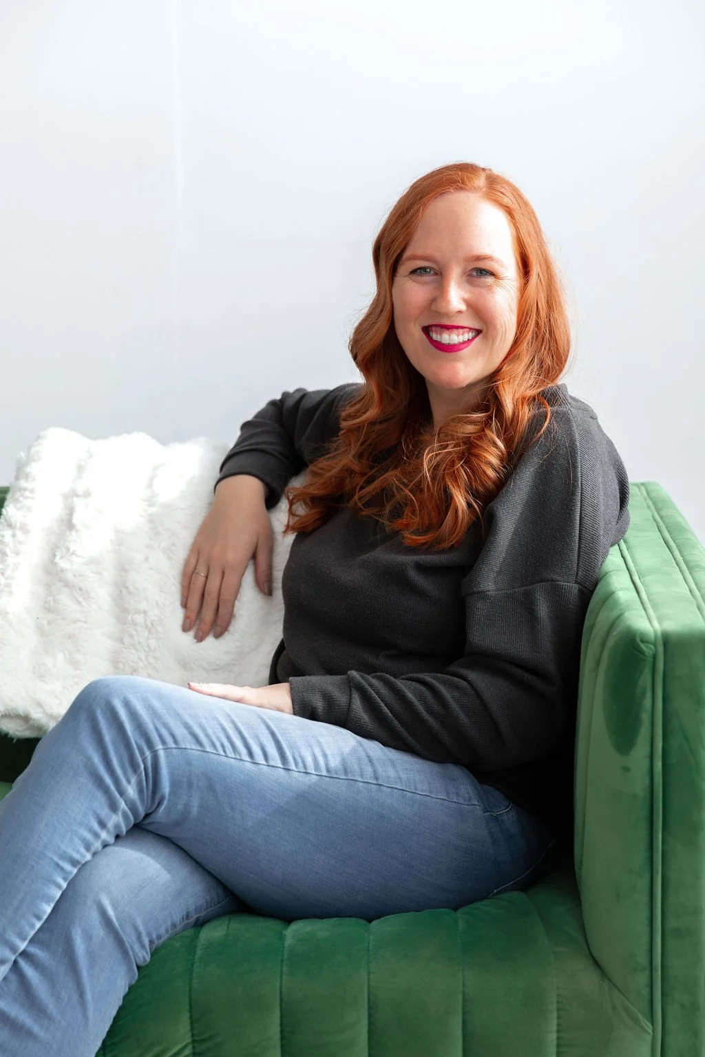 A woman with red hair and a gray sweater and jeans sitting on a green couch, smiling, with a white furry pillow behind her.