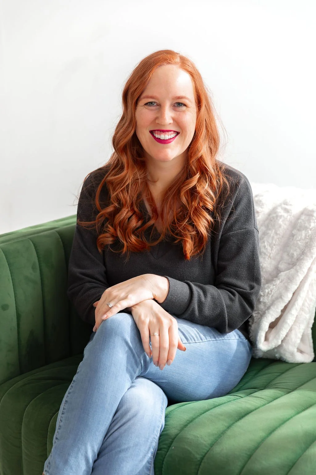 A woman with long red wavy hair smiling while sitting on a green sofa with her hands crossed.