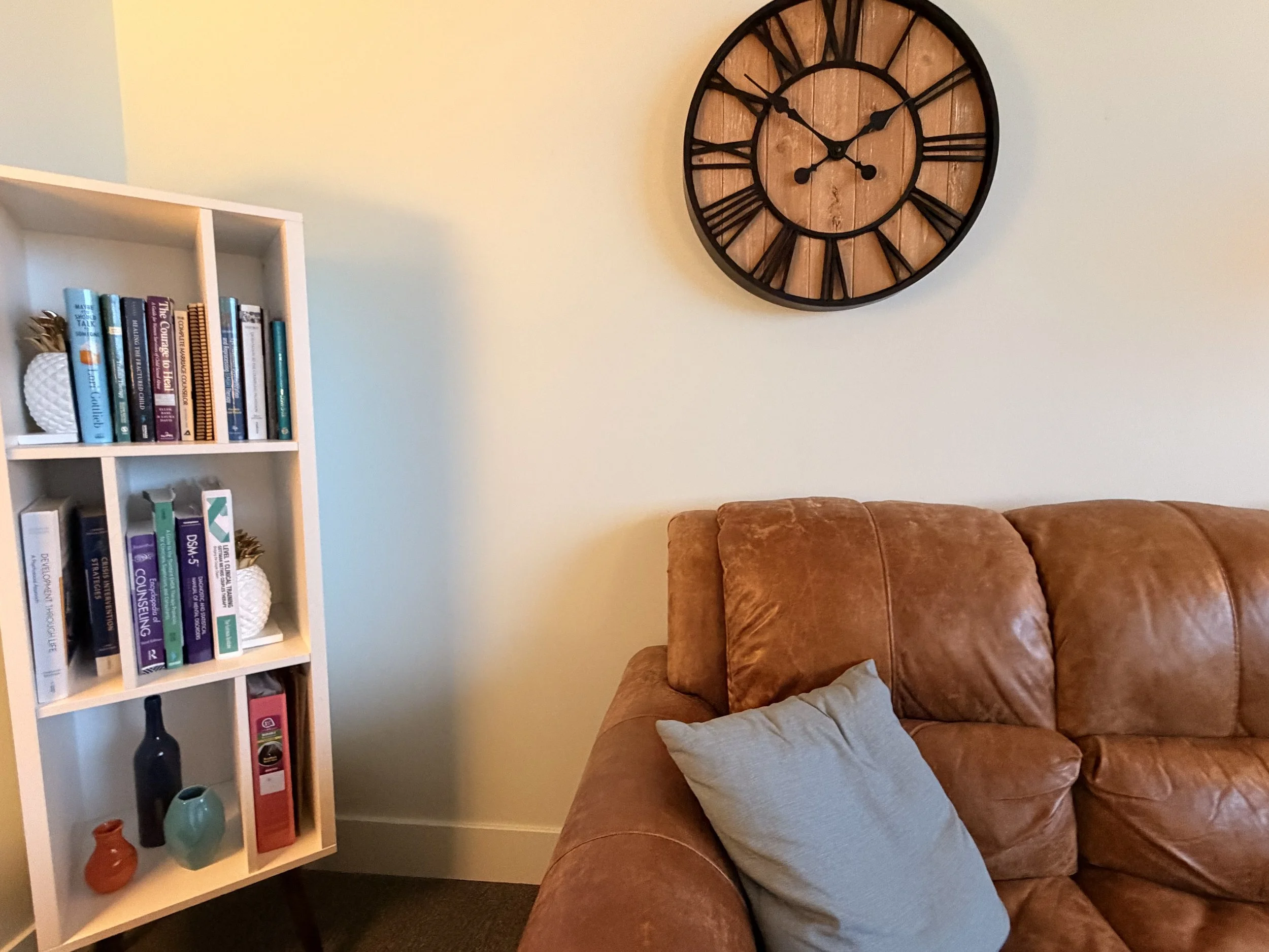 Cozy counseling office with white bookshelf filled with books and decorative vases, a brown leather sofa with a light gray pillow, and a large wooden wall clock with Roman numerals on a white wall.