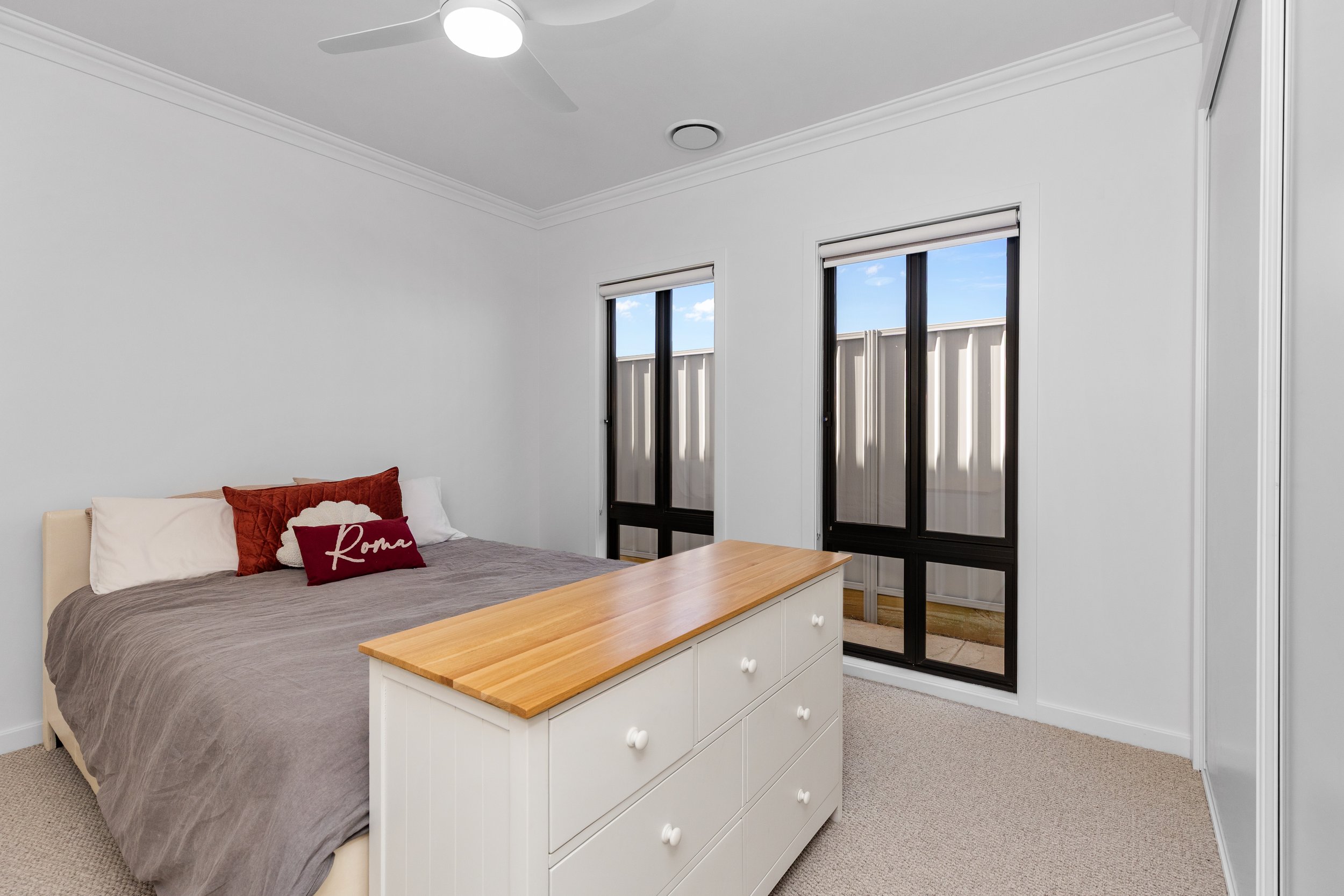 A bedroom with a bed, red and white pillows, a white dresser with a wooden top, and two large windows with black frames showing an outdoor fence and blue sky.