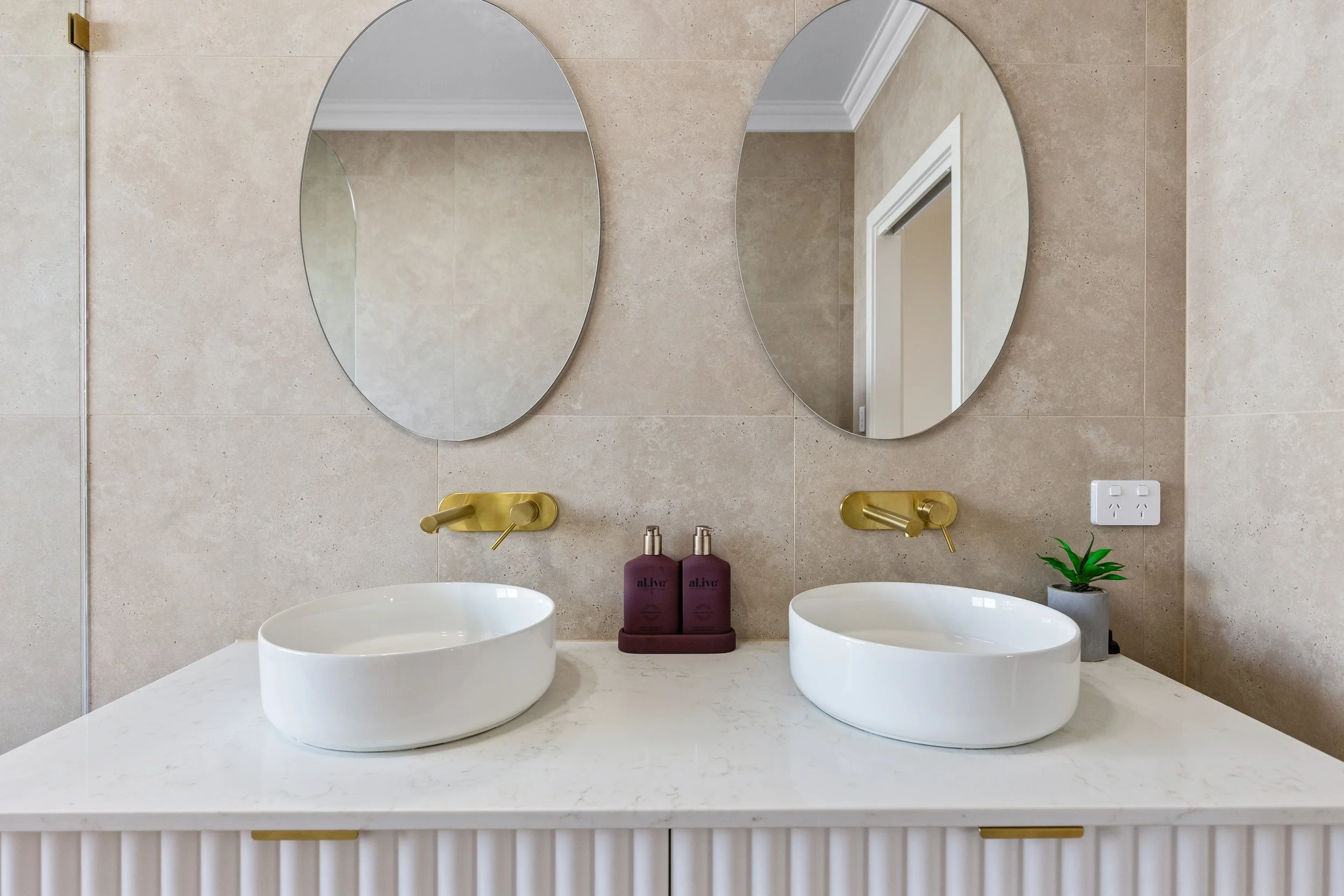 Bathroom countertop with two white vessel sinks, behind which are gold wall-mounted faucets, two purple soap bottles on a tray, a small potted plant, and a large mirror reflecting the beige-tiled wall and doorway.
