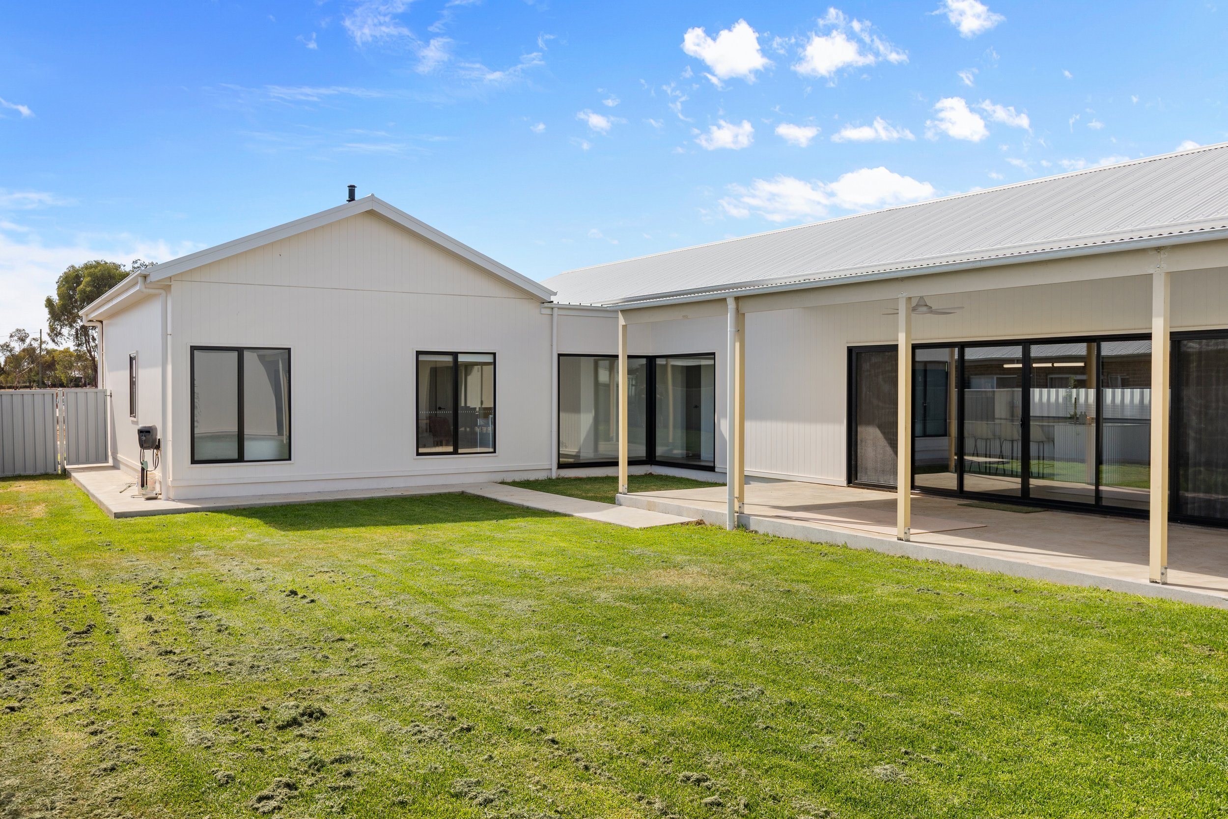 White house with a backyard, large glass sliding doors, and a covered patio with wooden posts, on a sunny day with blue sky and clouds.