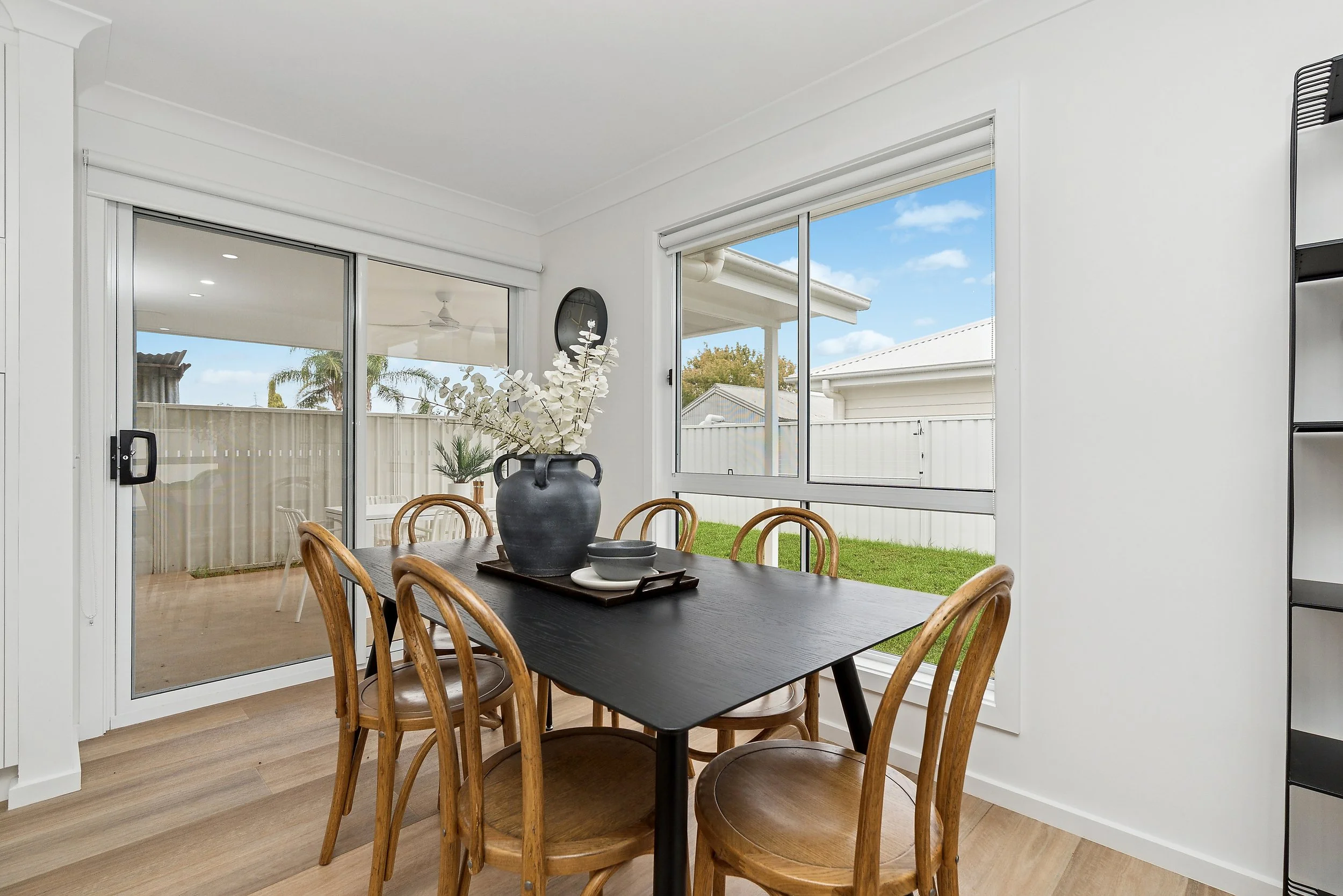 A dining room with a black rectangular table surrounded by six wooden chairs. The table has a black vase with white decorative branches, a tray with a bowl and cup. The room has large windows showing a backyard with grass and a white fence, and a sliding glass door leading to a patio with a ceiling fan.