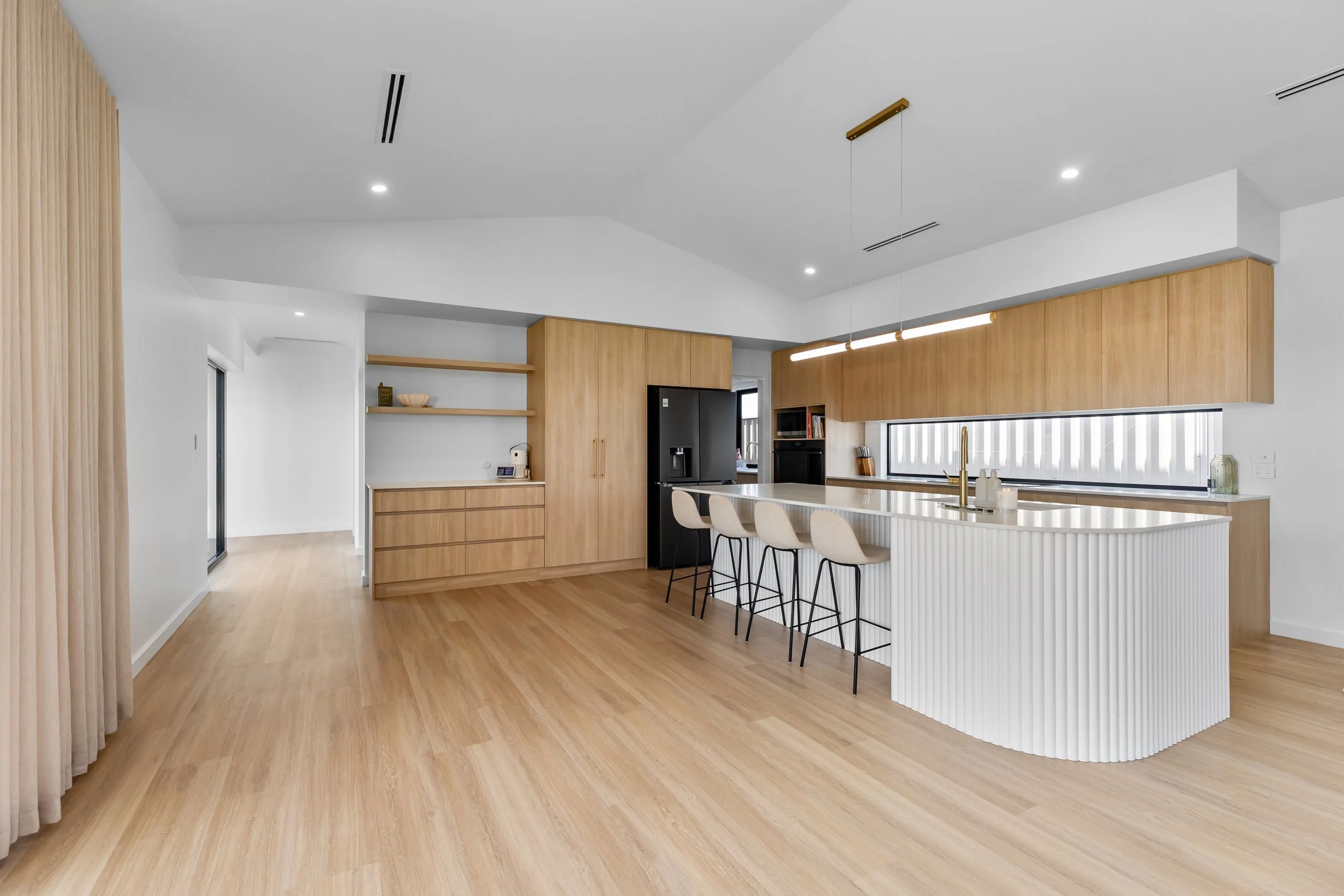 Modern kitchen with white and wood cabinetry, black refrigerator, island with white textured front and cream barstools, gold faucet, horizontal window shades, light wood flooring, and subtle ceiling lights.