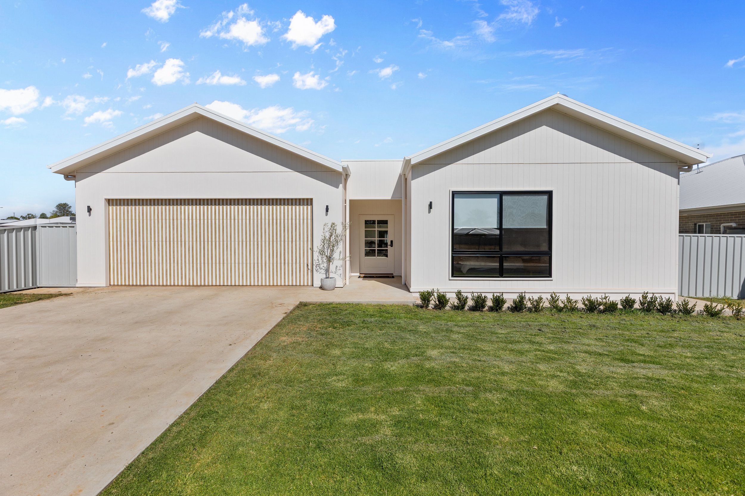 Modern white house with a two-car garage, black framed window, small front yard with green grass, and a concrete driveway, under a blue sky with a few clouds.