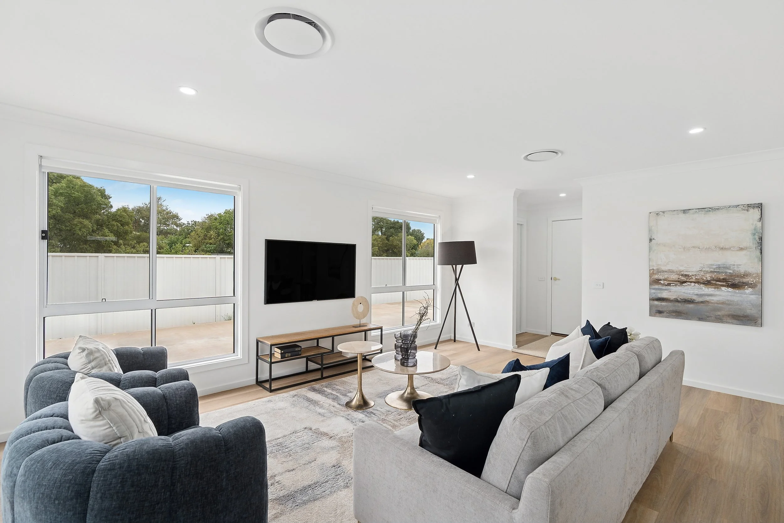 Sunlit modern living room with large windows, gray and black seating, a wall-mounted TV, a minimalist TV stand, a coffee table, a floor lamp, a beige rug, and abstract wall art.