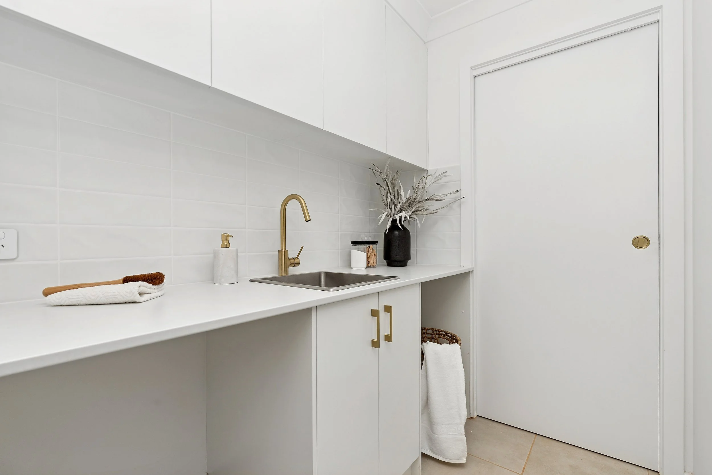 Minimalist white kitchen with a gold faucet, white countertop, and decor including a black vase with dried flowers, a soap dispenser, and a tray with a cloth and a wooden brush.