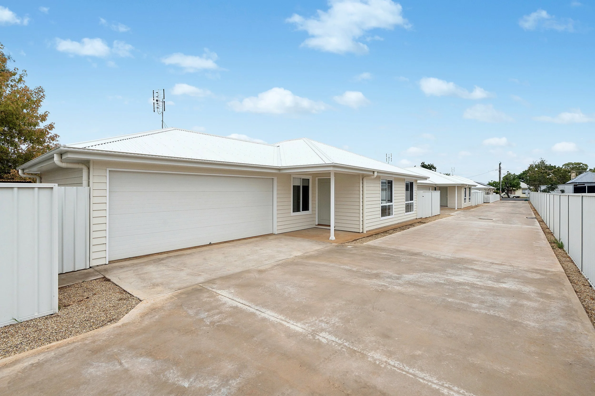 Single-story house with a white garage door, white vinyl siding, and a metal roof, surrounded by a concrete driveway and white fencing, under a partly cloudy sky.