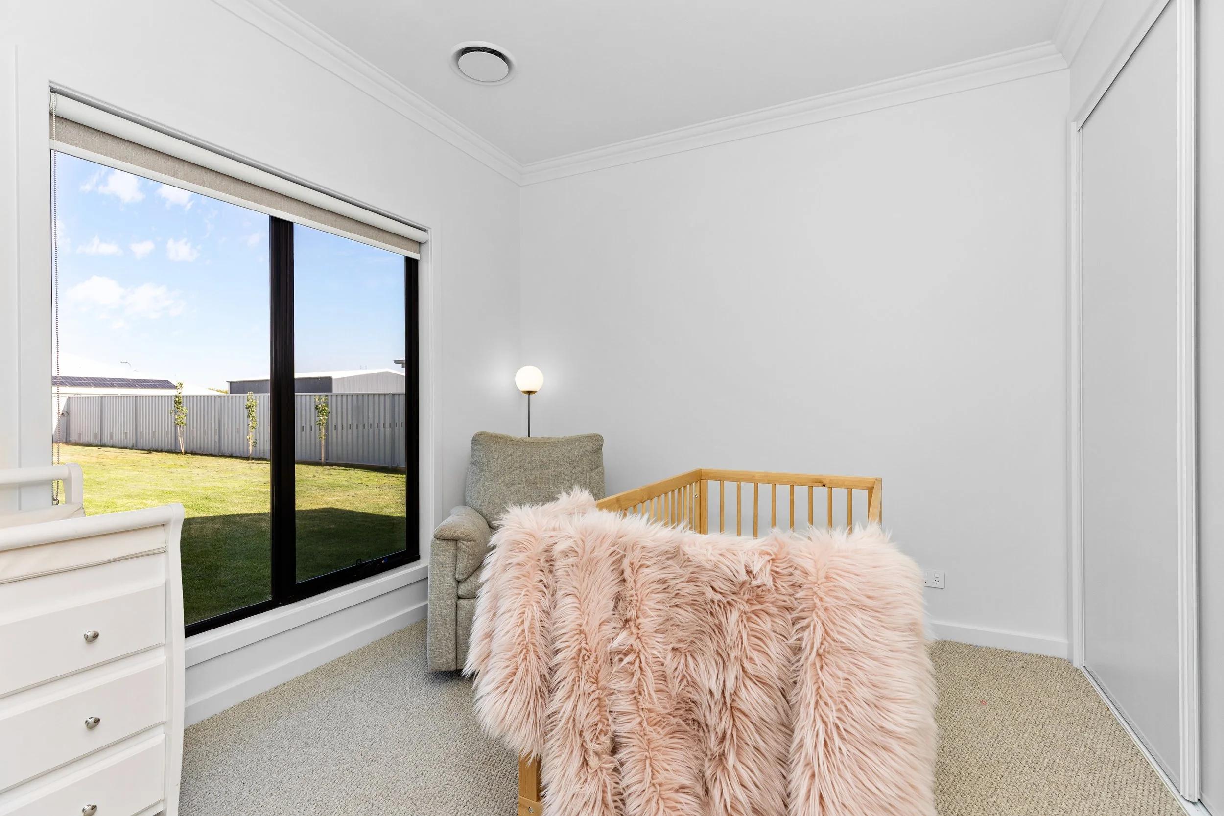 Nursery with white walls, beige carpet, large window with blinds showing backyard, beige armchair with lamp, wooden crib covered with pink fluffy blanket.