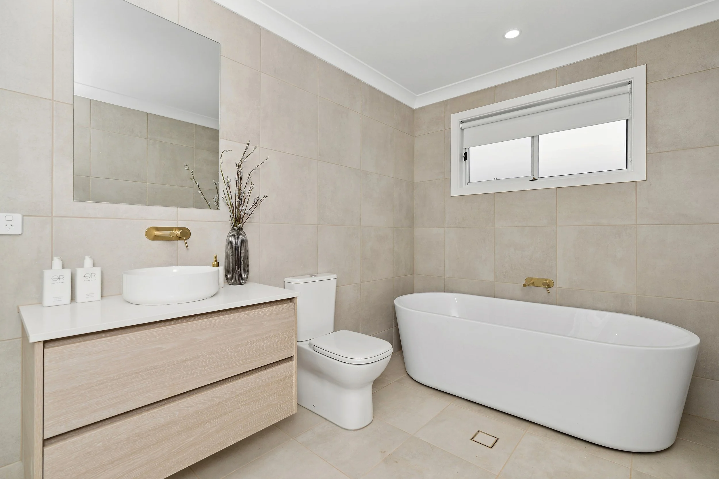 Modern bathroom with beige tiled walls and floor, a white bathtub, a toilet, a mirror above a wooden vanity with a white vessel sink, window, and decorative vase with branches.