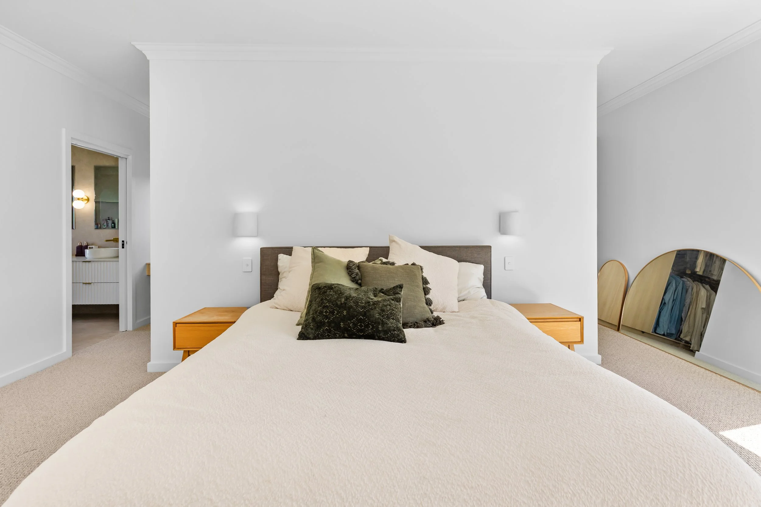 Neatly made bed with multiple pillows in a minimalist bedroom with white walls, nightstands, and a large mirror leaning against the wall.