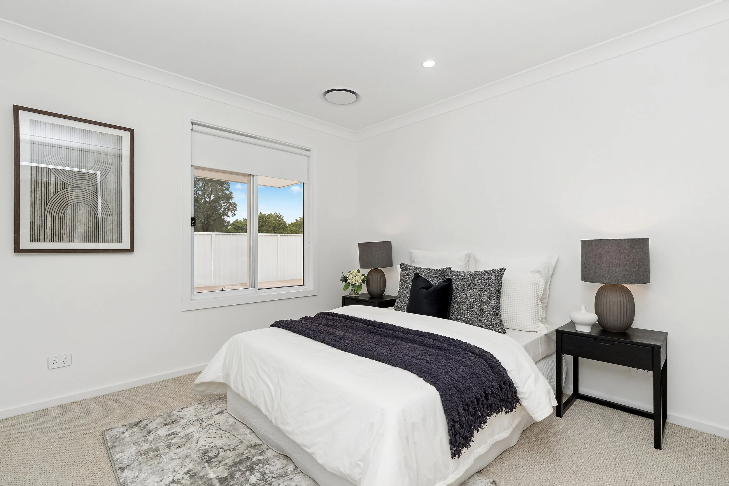 A minimalistic bedroom with white walls and beige carpet, featuring a bed with white bedding, black bedside tables with lamps, and modern framed artwork on the wall. A window shows an outdoor view of trees and a white fence.