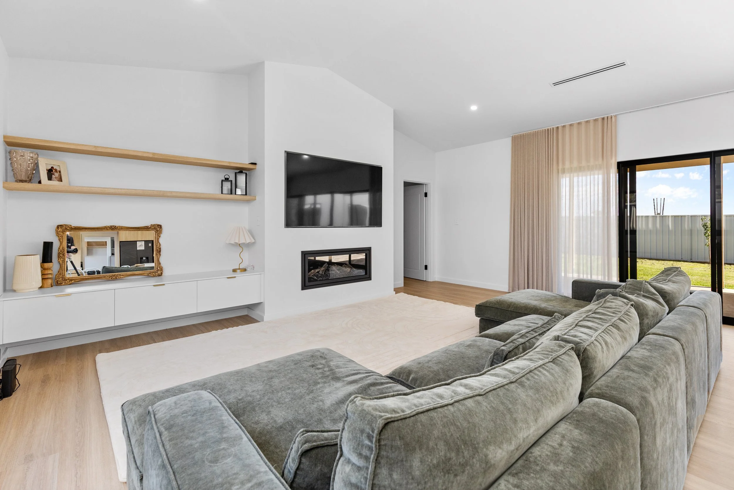 Modern living room with a gray sectional sofa, a white wall with a mounted TV and fireplace, open window with sliding glass door, and open in a well-lit space.
