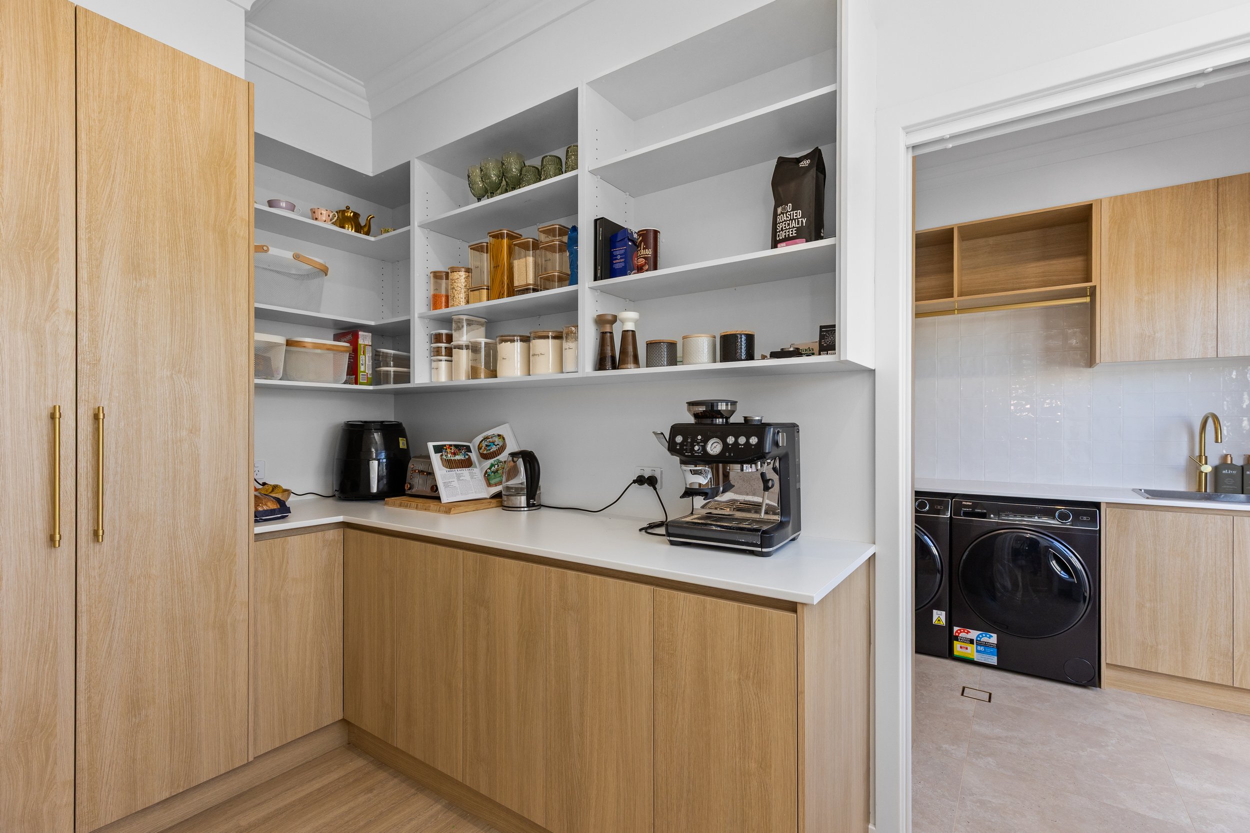 Kitchen with open white shelves displaying jars and containers, wooden cabinets, black coffee machine, air fryer, and an open doorway leading to a laundry area with black washer and dryer.