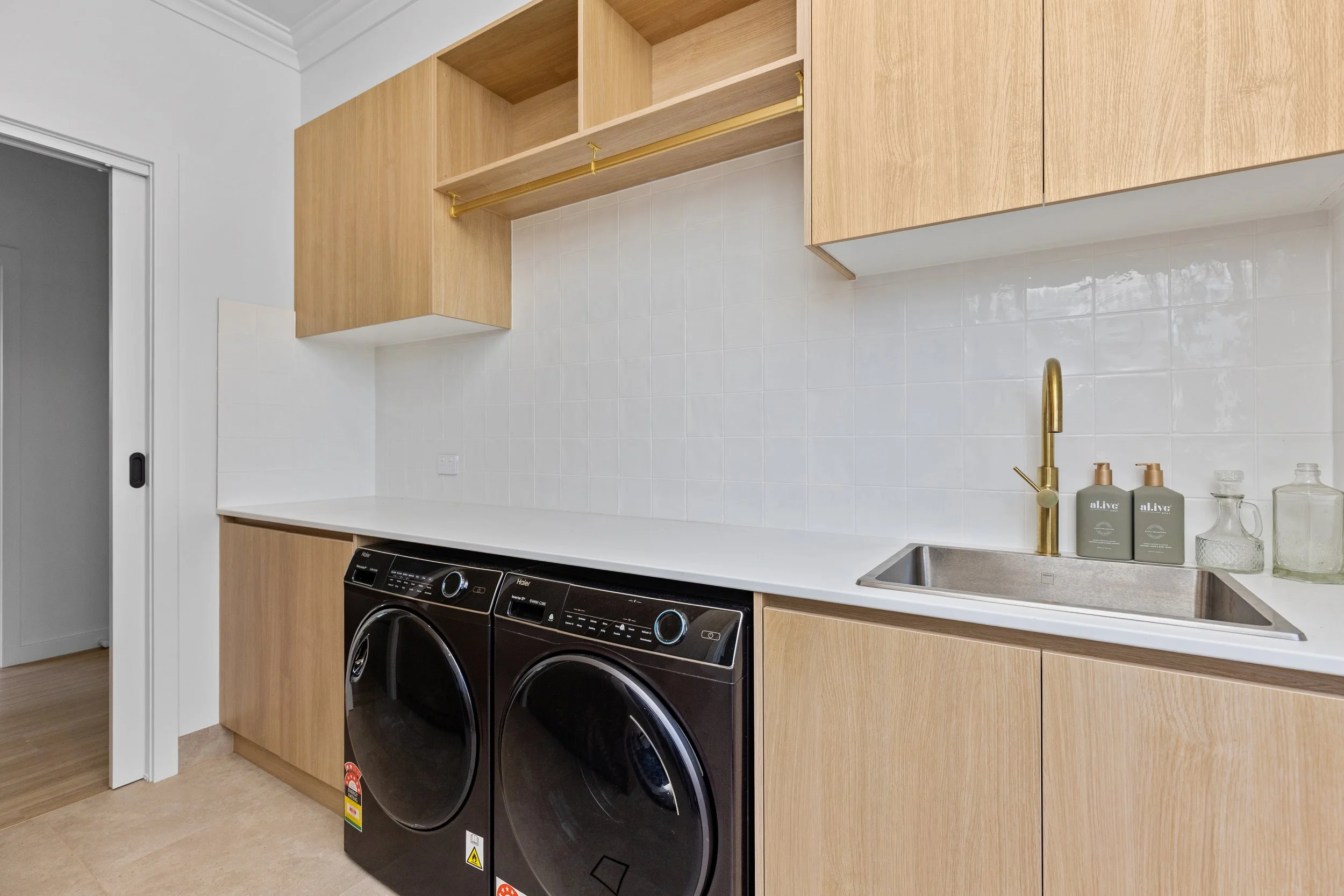 Laundry room with black front-loading washer and dryer, wooden cabinets, white countertop, and a sink with a gold faucet.