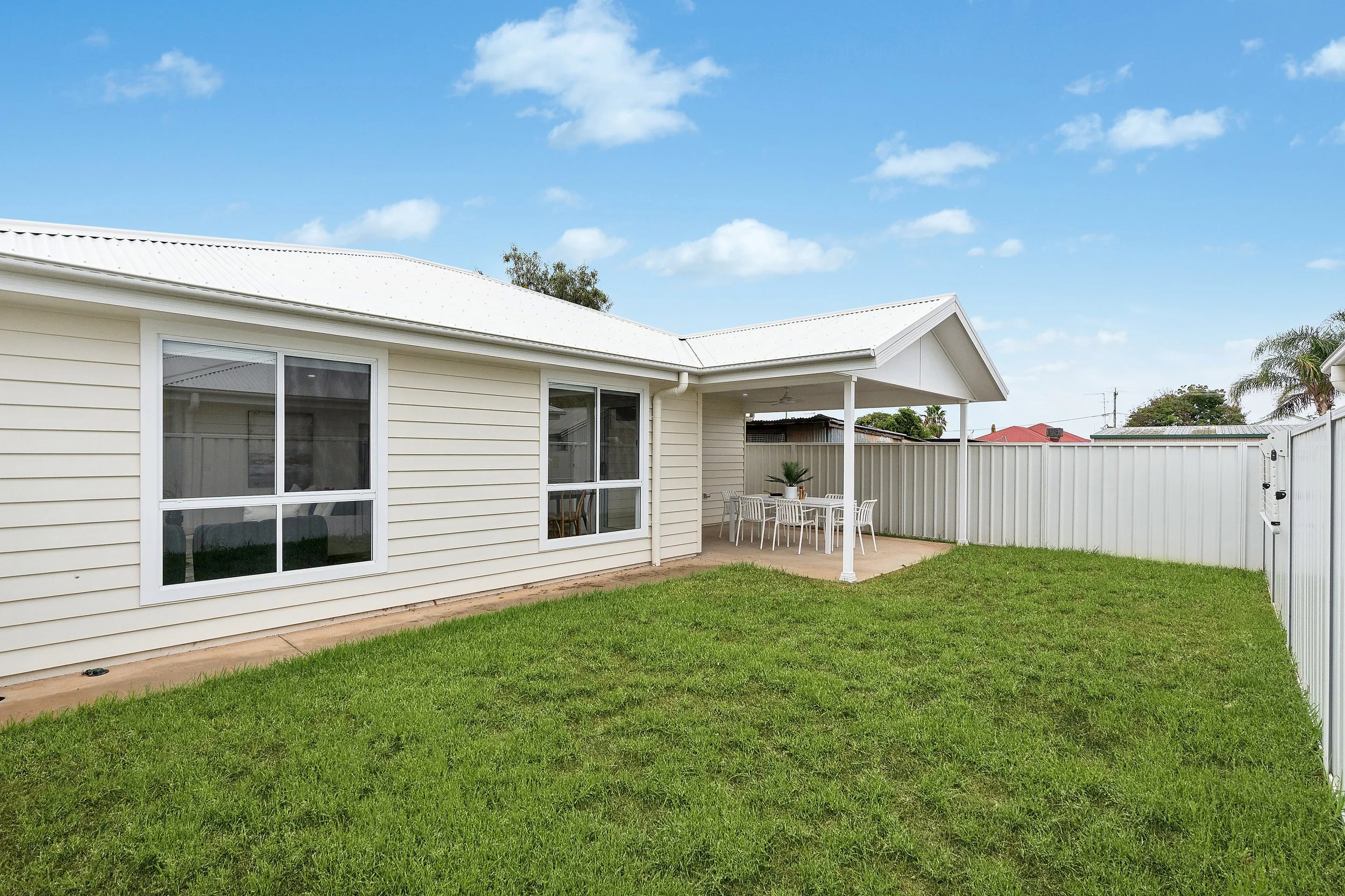 Backyard with green grass, white siding house with large windows, and a covered patio area with an outdoor dining table and chairs on a concrete slab, surrounded by a white fence under a partly cloudy blue sky.