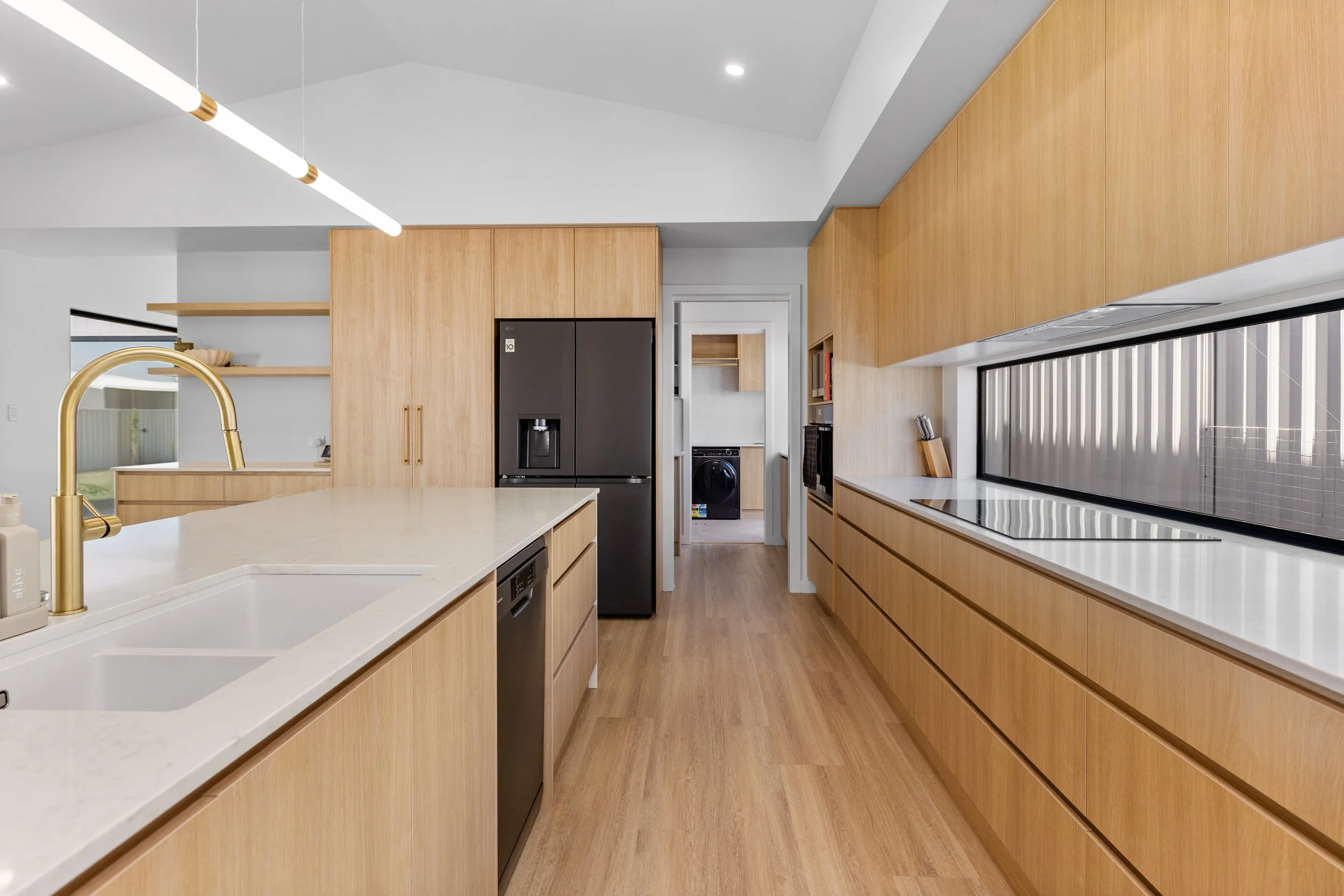 Modern kitchen with light wooden cabinets, white countertops, black appliances, and a gold faucet, with a view into a laundry area