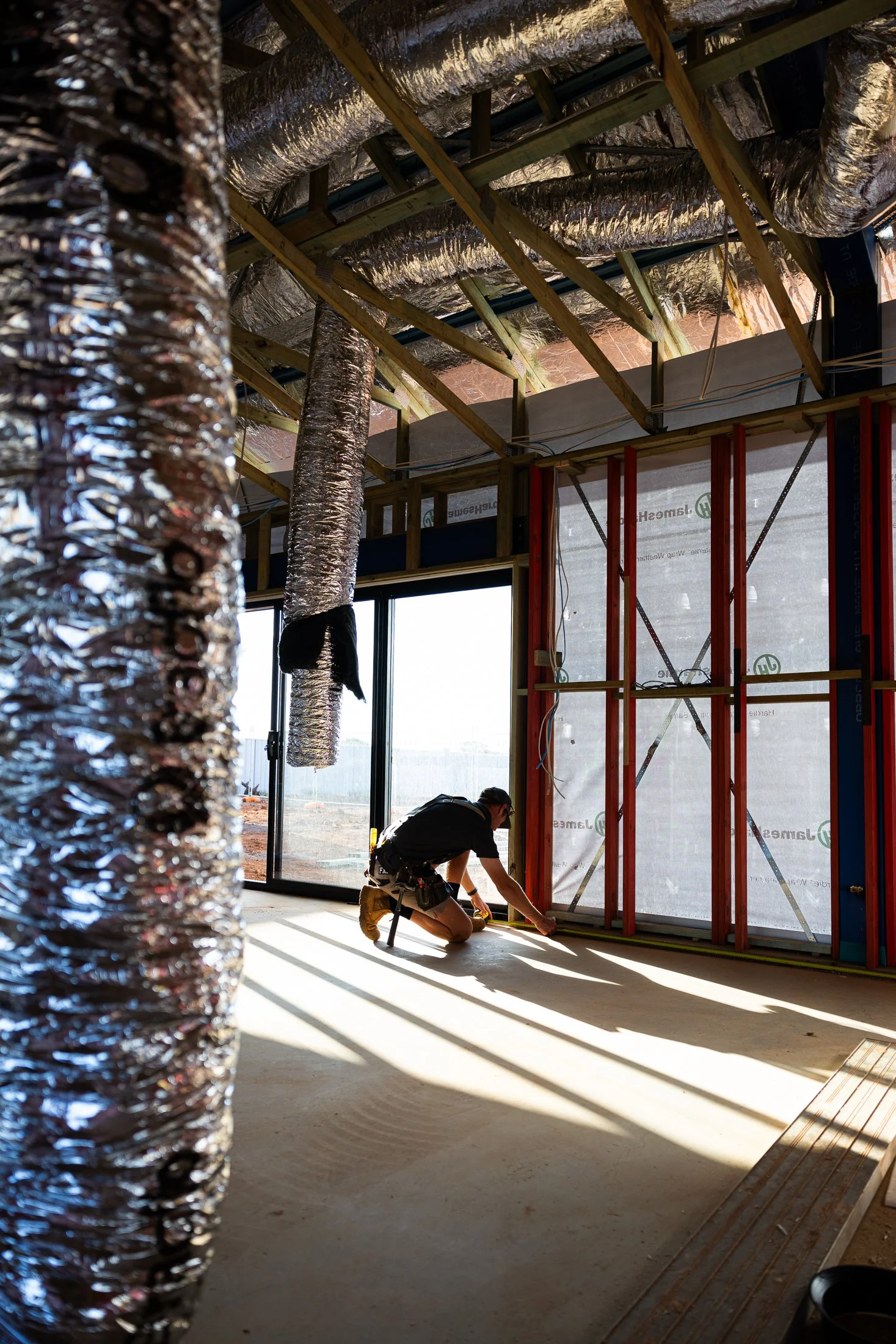 A construction worker kneeling on the floor inside a building under construction, measuring or marking a wall frame. Exposed ductwork is visible overhead, and large windows let in natural light.